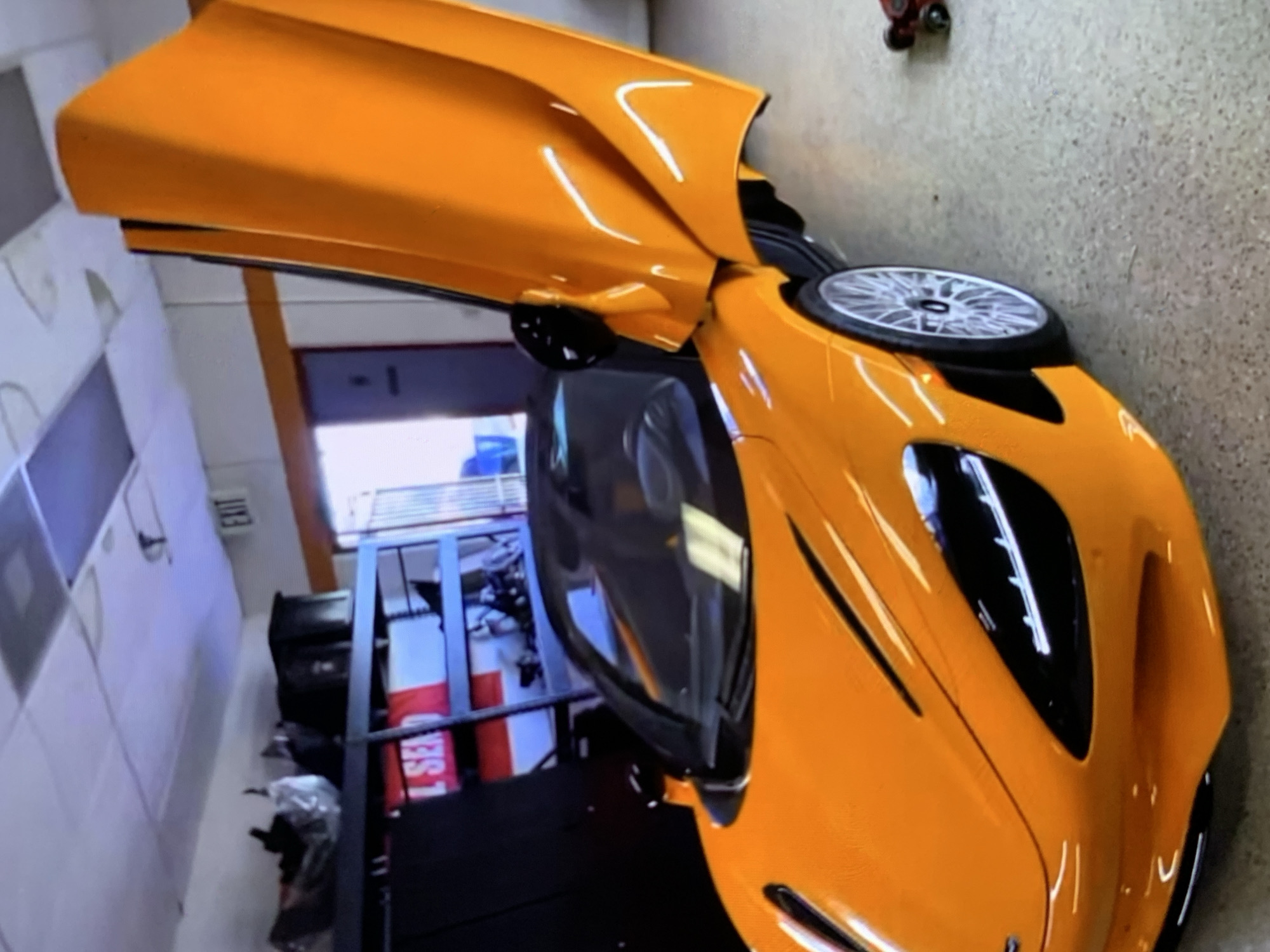 An orange McLaren supercar with dihedral doors open is parked indoors, possibly in a garage or workshop. The car is the primary subject, with its sleek, aerodynamic design and bright orange paint catching the light. The interior of the car is partially visible, showing black seats. The background includes shelves with various items, including boxes and what appears to be a red and white banner with "L SEA" printed on it. An "EXIT" sign is visible on the ceiling. The flooring is a speckled gray material, and a small red object, possibly a toy car or tool, is on the floor to the right of the vehicle. The lighting suggests an indoor environment, with fluorescent lights visible on the ceiling.