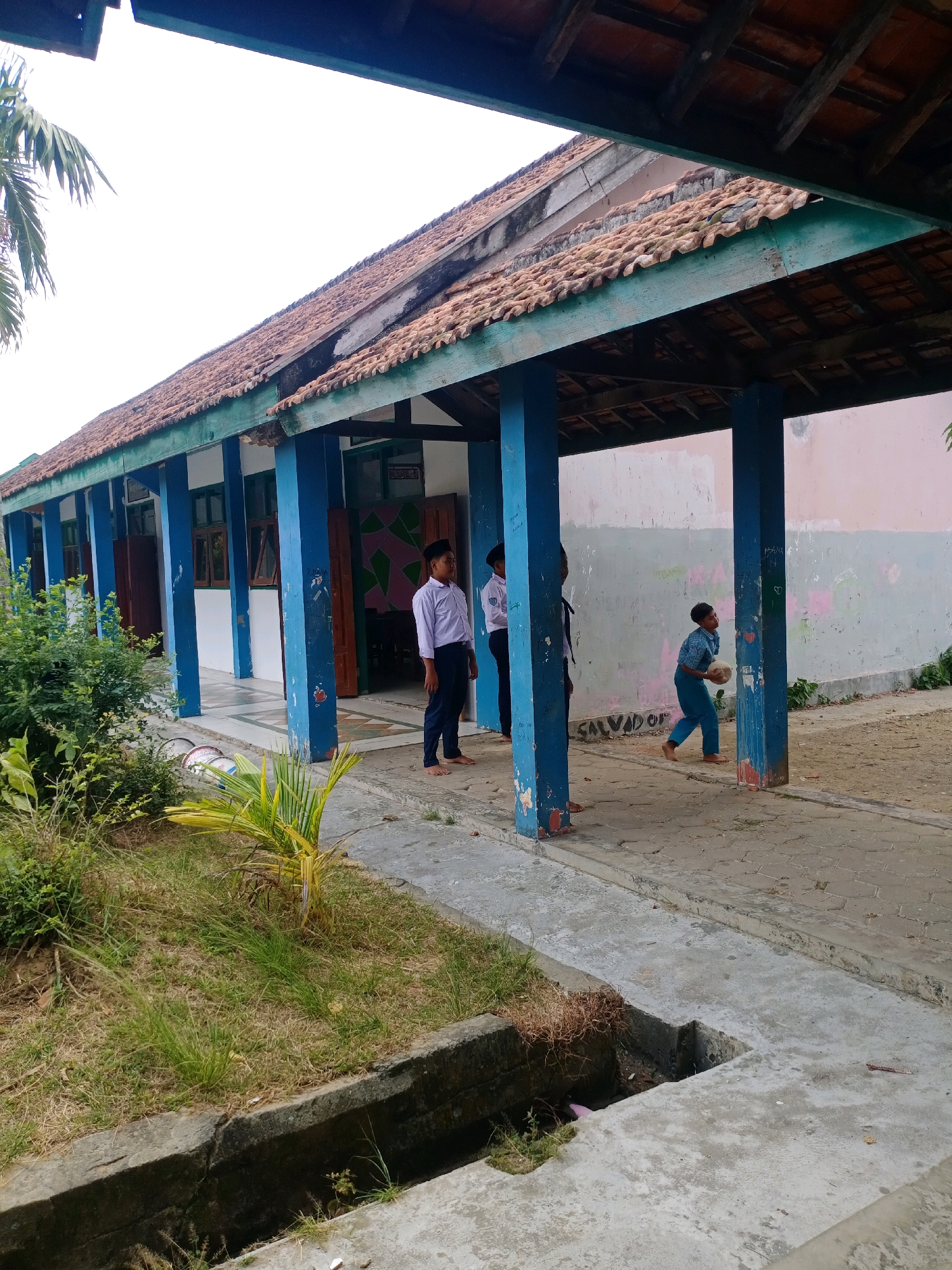 The image depicts an outdoor scene at a school building in Tragah, Indonesia. A covered walkway with blue pillars and a tiled roof runs along the side of a building with multiple doors and windows. Three young boys are present. One boy, wearing a white shirt and dark pants, stands near the left side of the walkway. Another boy, in a similar uniform, stands further down the walkway. A third boy, wearing a blue shirt and pants, is in the process of throwing a round, light-colored object, possibly a ball. The ground is paved with concrete, and there is a grassy area with a drainage ditch to the left of the walkway. The walls of the building are painted in muted colors and show some graffiti. Vegetation, including a palm frond, is visible to the left of the walkway. The sky is overcast.