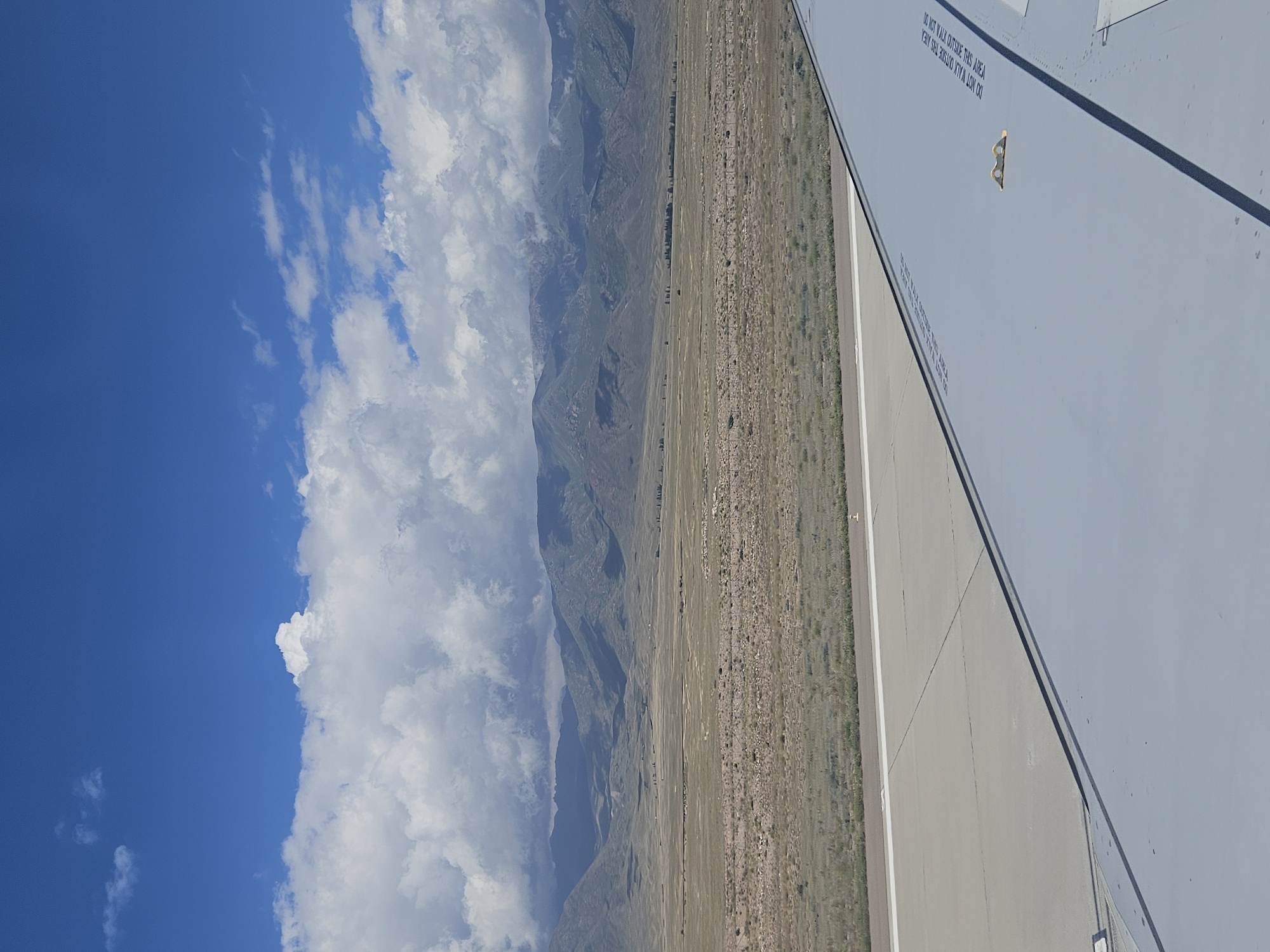 The image captures a view from an airplane wing, looking out towards a mountainous landscape under a partly cloudy sky. The foreground features the gray surface of an airplane wing with visible warning text and a yellow lifting point. Adjacent to the wing is a tarmac runway with white markings. Beyond the runway, a vast, arid plain stretches towards rolling, green-covered mountains. The sky above is a bright blue, dotted with fluffy white clouds that cast shadows on the distant peaks. The scene is likely the airport in Tamchy, Kyrgyzstan, given the distinctive landscape often seen in that region. There are no people visible in the image.