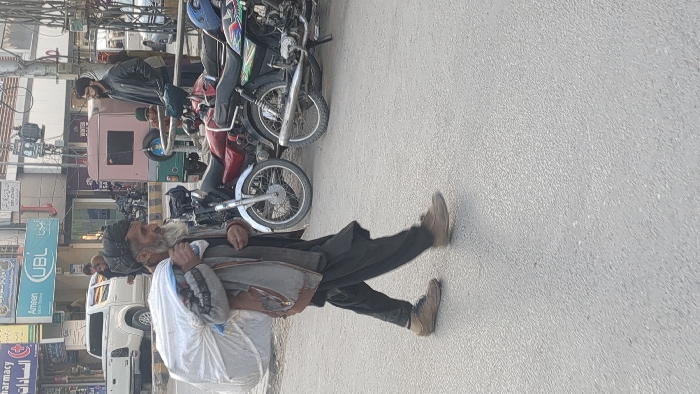 This image depicts a daytime urban street scene in Quetta City Tehsil, Pakistan. The primary subject is an elderly male with a long white beard and a dark cap, walking from left to right on a paved road. He is dressed in a grey-brown jacket over dark attire and light brown shoes. He carries a large white sack over his left shoulder, supported by his left hand, and a small clear plastic bottle in his right hand.
In the middle ground, several motorcycles are parked, including models in red and green colors. Behind these, a light pink auto-rickshaw is visible. A young male with a dark beard and a dark jacket stands near the auto-rickshaw, oriented towards the left. A smaller individual in a dark cap is partially obscured nearby.
The background consists of commercial buildings. Prominent signage includes "UBL" and "Ameen Islamic Banking," displayed in both English and Urdu, alongside a "Pharmacy" sign marked with a red cross. A white pickup truck is parked on the left side of the street. Utility poles with associated wiring are also present.
