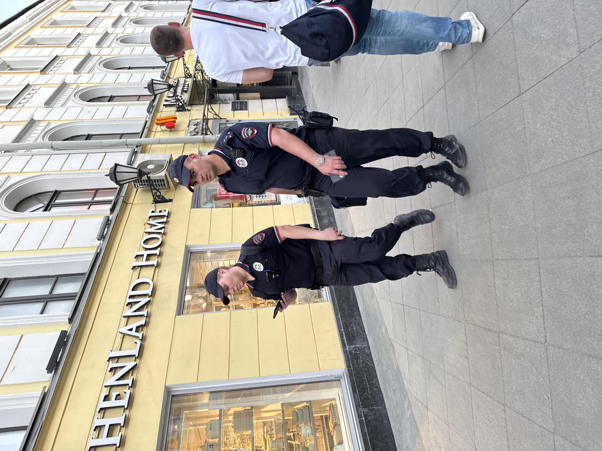 Two Russian police officers are standing on a sidewalk in Moscow, Russia. They are both wearing dark blue uniforms and black boots. One is holding a smartphone. Behind them is a yellow building with the sign "HENLAND HOME" vertically aligned. An overhead sign reads, "МЕНЮ ВКУСКИЙ". A person with a white shirt and blue jeans is walking toward the officers.