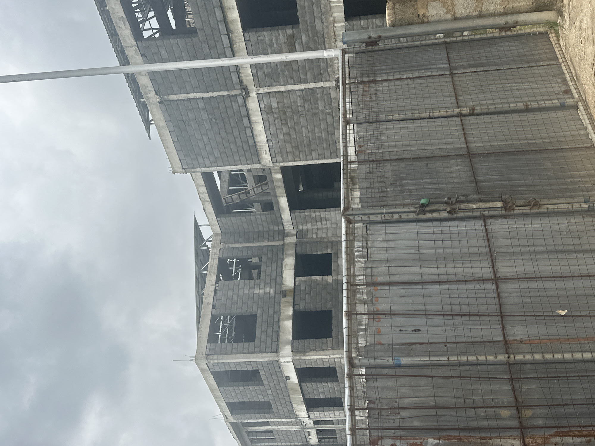 This is a photograph of a multi-story building under construction in Gwarinpa, Nigeria. The structure appears to be made of concrete blocks and has exposed rebar and unfinished roofing. In the foreground, a metal security fence with a wire mesh covers the entrance to the construction site. The sky is overcast, suggesting a cloudy day. The scene is devoid of people.
