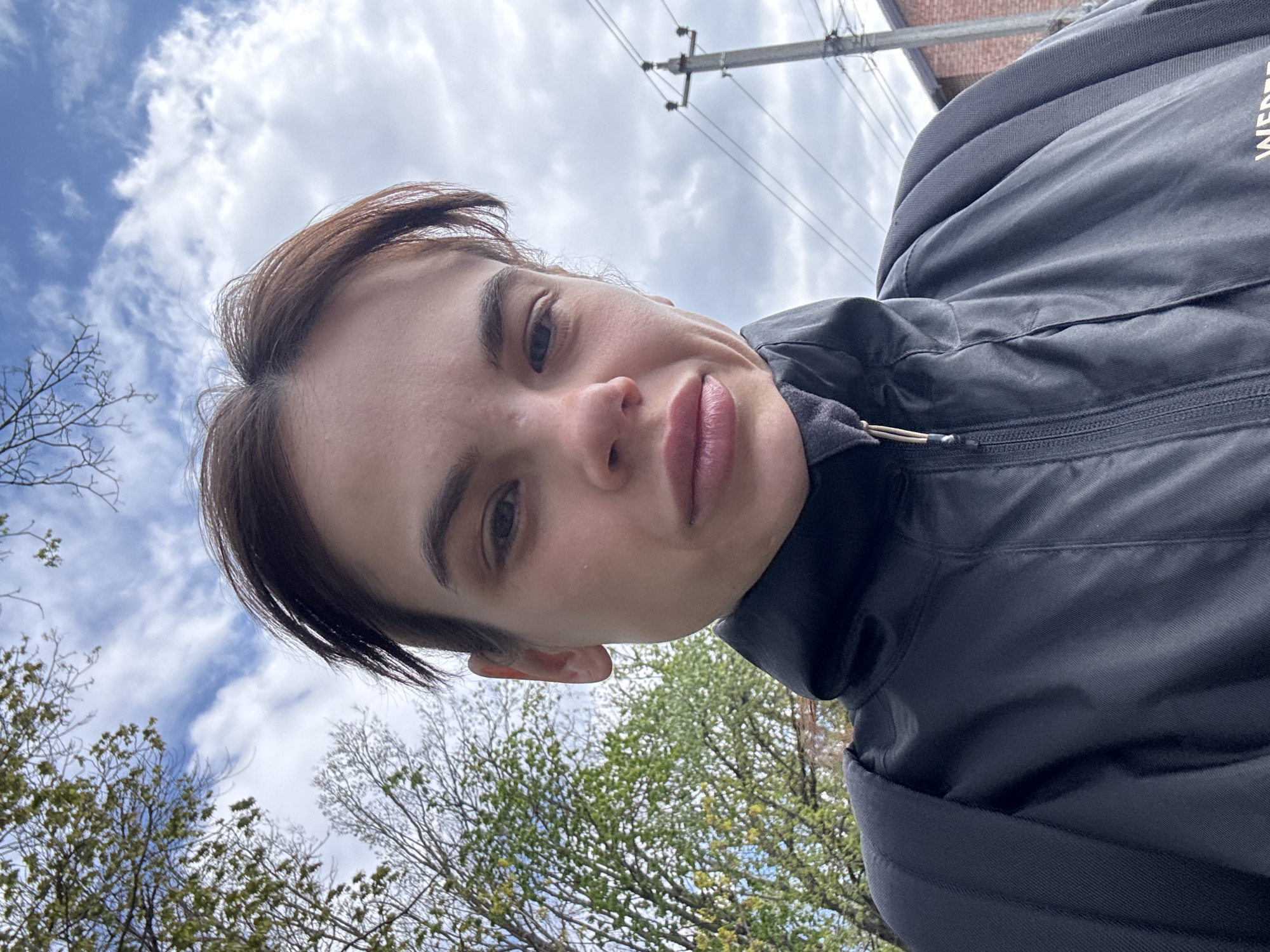 The image is a low-angle, rotated selfie portrait of a young person, likely female, with short dark hair, captured from below looking upwards. The person gazes directly at the camera with a neutral to slightly pouting expression, their head tilted. They are wearing a dark grey or black zippered jacket, with a partially visible white logo or text, possibly starting with "WED," on the right side.
The background suggests an outdoor urban or suburban setting in Rostov-on-Don, Russia. The sky is largely cloudy with patches of blue visible. In the upper right quadrant, utility poles and overhead power lines traverse the sky, and a reddish-brown brick building can be seen in the distance. The lower part of the image features trees with green leaves, indicating spring or early summer, alongside some bare branches on the far left. The lighting is bright and diffused due to the overcast sky, suggesting it is daytime. No specific activity beyond taking the selfie is depicted.
