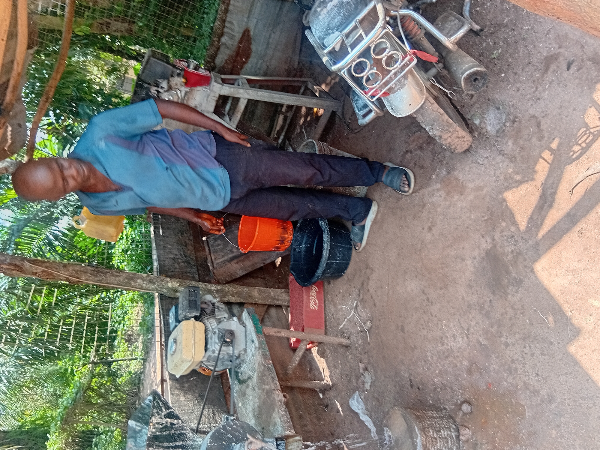 A bald, dark-skinned man is standing in a dirt-floored outdoor area, possibly a workshop or a yard, in Ejule, Nigeria. He is wearing a blue short-sleeved collared shirt and dark trousers. He is holding an orange bucket in his right hand. To his left, there is a generator on a table, an orange Coca-Cola crate, and a black tub. To his right, a motorcycle is propped up, and a metal rack is visible behind it. The background consists of lush green foliage and trees. The lighting suggests it is daytime.