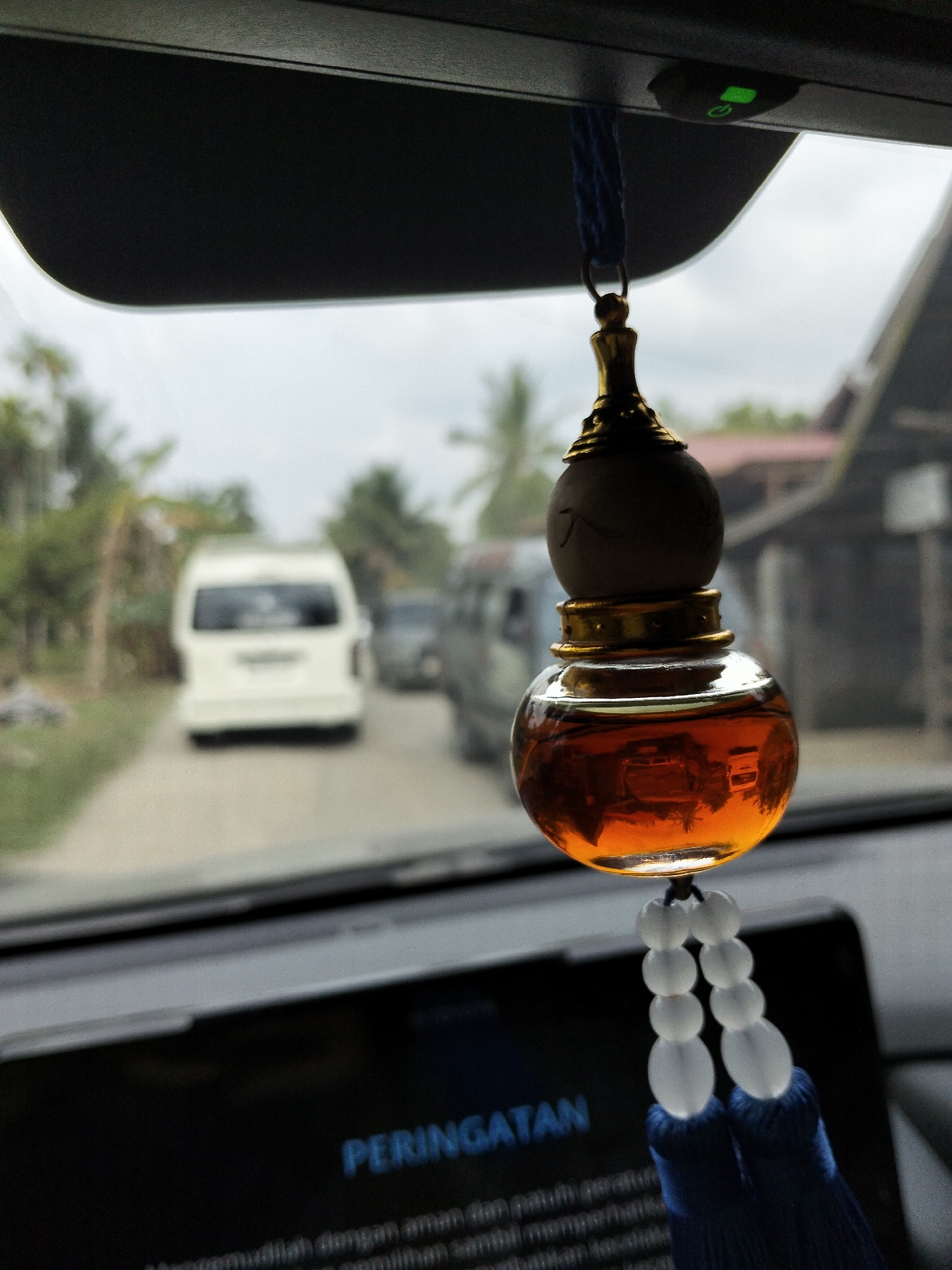 This is a close-up, indoor shot from inside a vehicle, looking forward. The primary subject is an amber-colored liquid in a decorative, gourd-shaped glass bottle hanging from the rearview mirror. The bottle has a gold-colored cap and base, and is adorned with a string of white beads and a dark blue tassel. In the background, the scene is slightly out of focus, showing a road with two white vans, and possibly other vehicles. Trees and buildings are visible on either side of the road, suggesting a rural or semi-rural setting. The sky appears overcast, indicating a cloudy day.
The text "PERINGATAN" is visible on a dark screen in the foreground, likely a car's dashboard display. This translates to "WARNING" in Indonesian. The lower part of the screen shows more text, also in Indonesian, which appears to be a safety message related to driving. The overall impression is one of being a passenger in a vehicle, possibly in a tropical or subtropical region, given the vegetation. The image evokes a sense of everyday travel. The lighting suggests it is daytime.