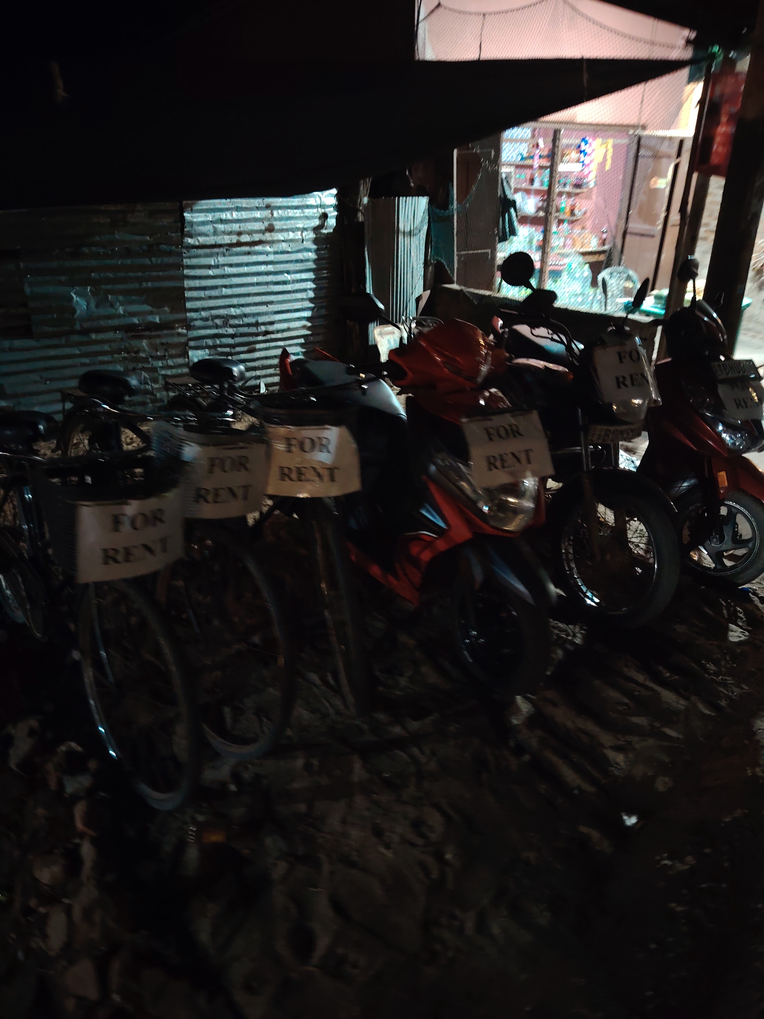 The image shows a row of parked motorbikes and bicycles outside a small shop at night. The shop appears to be a general store or a small convenience store, with shelves of items visible through the window. The vehicles, mostly motorbikes, are lined up in front of a corrugated metal wall. Several of the motorbikes have "FOR RENT" signs attached to them, indicating they are available for hire. Some bicycles are also visible, also with "FOR RENT" signs. The scene is dimly lit by artificial light, suggesting it is nighttime. The ground appears to be dirt or gravel, and the overall setting is rustic and somewhat dilapidated. The mood is quiet and still, with no people visible in the immediate foreground. The focus is on the vehicles and the shop, creating a sense of a business that operates at this location. The presence of the rental signs suggests a tourist area or a place where temporary transportation is commonly needed.