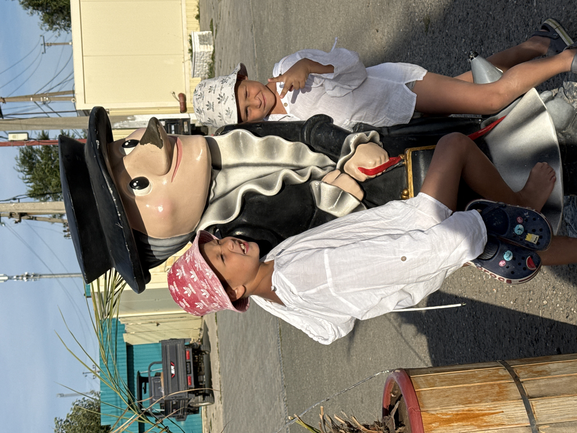 Two young boys are posing with a life-sized statue of Pinocchio in Sol-Iletsk, Russia. The boy on the left, wearing a red bucket hat and a white tunic, leans against the statue. The boy on the right, wearing a white bucket hat and a white tunic, stands next to the statue, making a peace sign with his hand. The Pinocchio statue is dressed in a black hat and coat. The background shows a street scene with buildings, utility poles, and a small truck. The image appears to be taken on a sunny day.