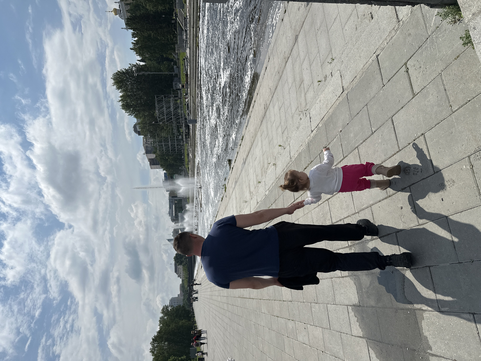 In Yekaterinburg, Russia, on a sunny day with scattered clouds, a man and a young girl walk hand-in-hand along a wide, paved walkway. The man, wearing a dark blue t-shirt and black pants, carries a dark jacket in his left hand. He is walking away from the camera, his right hand holding the girl's left. The girl, with blonde hair in a ponytail, wears a white long-sleeved shirt and pink pants. She is also walking away from the camera, her right hand extended slightly forward as if reaching for something.
The walkway is made of large, light grey rectangular pavers. To the right of the walkway is a wide, flowing body of water, possibly a canal or river, with numerous fountains spraying water into the air. The water surface is choppy and reflects the sunlight. Beyond the water, on the opposite bank, is a park with mature trees. In the background, the Yekaterinburg city skyline is visible, featuring modern buildings and some older structures, including a building with a golden dome. A large metal scaffolding structure is also present near the treeline. The overall atmosphere suggests a recreational area in the city.