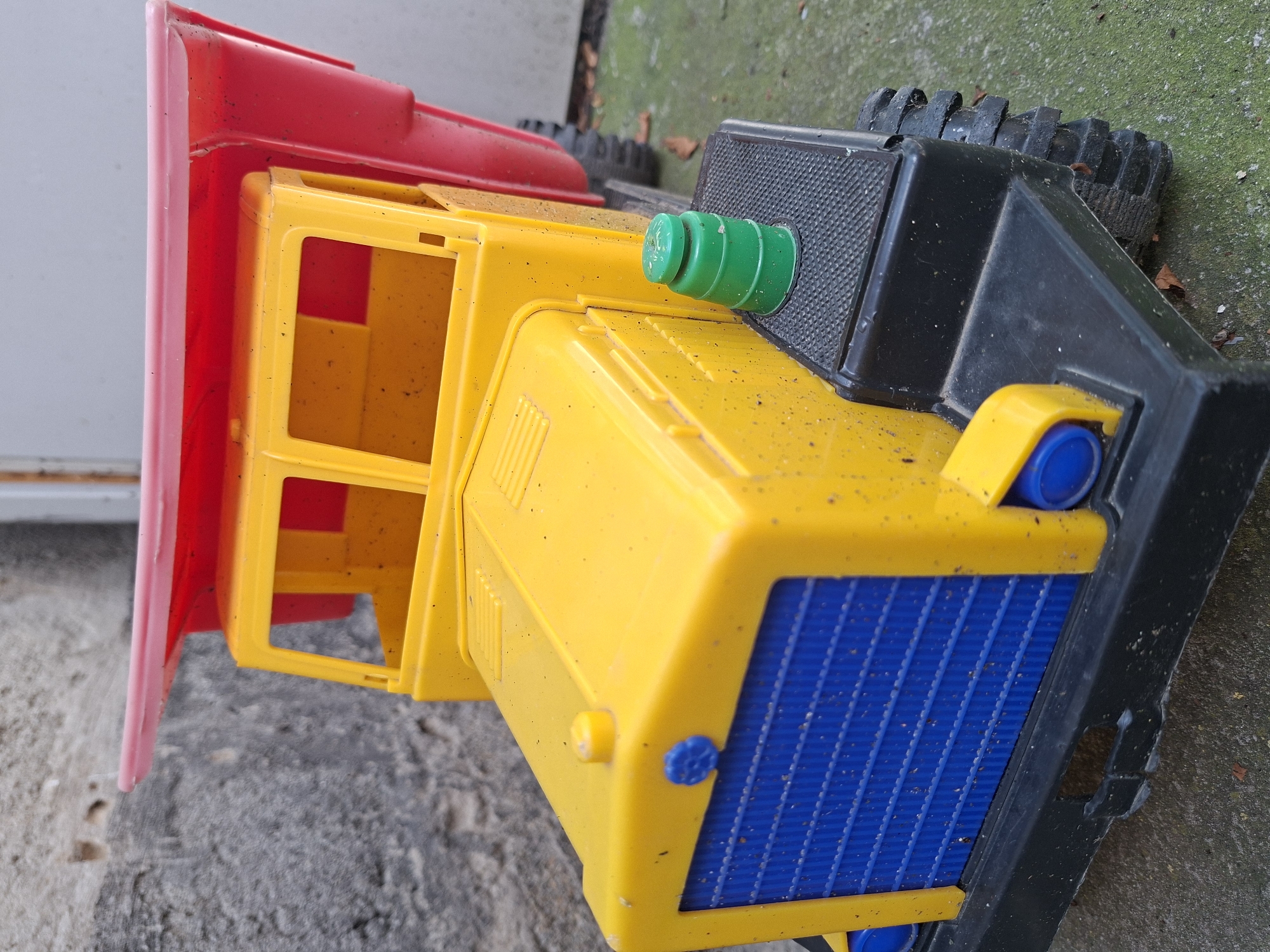 A close-up, eye-level shot focuses on a yellow, red, and blue plastic toy dump truck parked on a mottled gray and green surface, possibly concrete or asphalt. The truck's front features a blue grille and a yellow hood with a blue circular emblem and a yellow cap. A green barrel-shaped cap sits on the truck's black step. The truck's red bed is raised slightly. The background consists of a textured gray wall with some discoloration. The scene is outdoors, with scattered dry leaves on the ground. The lighting is natural, creating shadows and highlights on the truck's plastic surfaces. No people are present. The image does not provide any specific information about the location within Gorkovskiy, Russia.