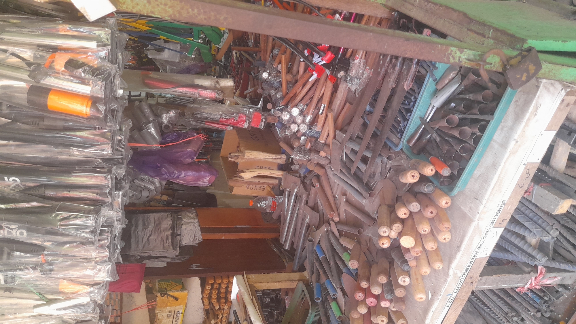 This is a full, eye-level shot of a tool stall in Pekalongan, Indonesia. The stall is packed with various tools, primarily agricultural and construction implements, displayed both hanging and piled on the ground. Hanging from the ceiling are numerous tools, mostly hoes and machetes, encased in clear plastic. In the foreground, a collection of wooden-handled tools, including hoes and shovels, are neatly arranged on a wooden counter. To the right, a green plastic container holds several metal tools with conical ends, possibly augers or drills. A rusty padlock hangs from a green metal beam on the right side of the frame. The stall appears to be outdoors or in an open-air market, with a partially visible wooden structure supporting the display. The overall impression is one of a busy marketplace with a wide variety of tools for sale.