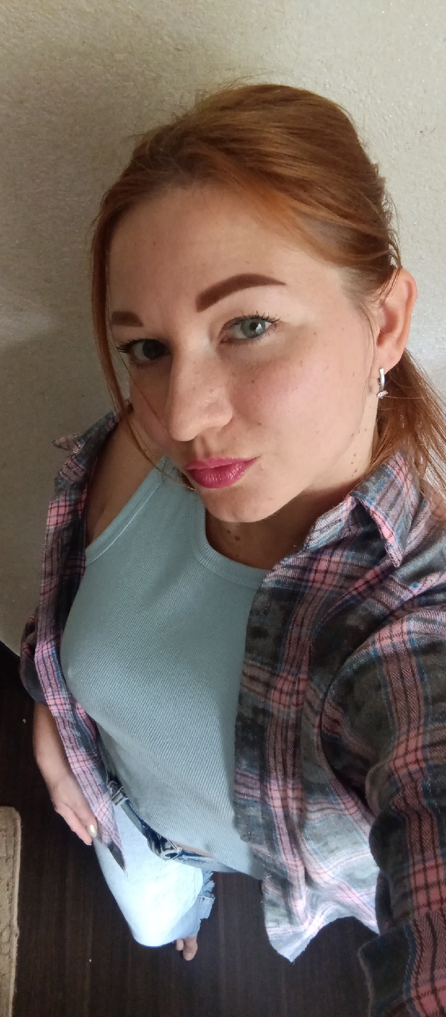 A high-angle shot depicts a woman with red hair, wearing a light blue tank top and a pink and grey plaid shirt. She has fair skin with freckles, light eyes, and is wearing pink lipstick. She also appears to be wearing an earring. The background is a neutral-colored wall and dark flooring. Parts of denim jeans are visible on her lower half. Her location and specific environment within the city are unknown.