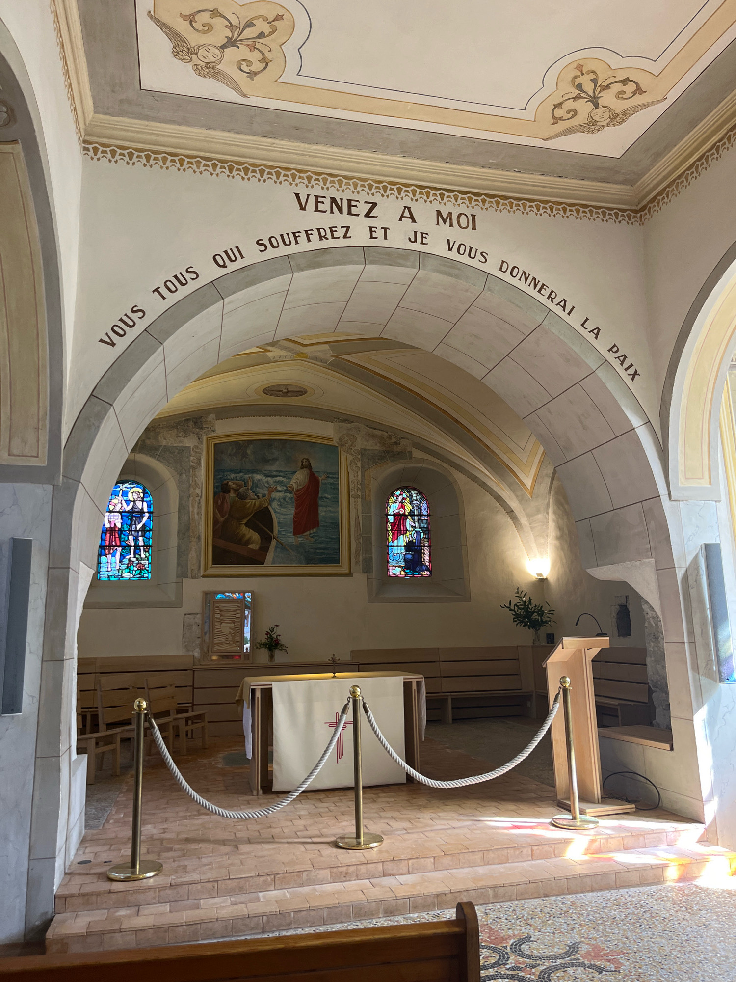 The image shows the interior of a chapel in Yvoire, France. The chapel features an arched ceiling with decorative frescoes and painted borders. Arching over the central altar area is an inscription in French that reads "VENEZ A MOI VOUS TOUS QUI SOUFFREZ ET JE VOUS DONNERAI LA PAIX," which translates to "Come to me, all who are weary, and I will give you rest."
In the foreground, a low barrier rope with brass stanchions separates the viewer from the altar area. The altar is covered with a white cloth, and a red cross design is visible. Behind the altar, there are rows of wooden pews. To the left of the altar, a stained-glass window depicts religious figures. To the right, a wooden lectern stands illuminated by sunlight streaming from a window. A large painting of Jesus walking on water hangs on the wall behind the altar. The overall environment is serene and religious.