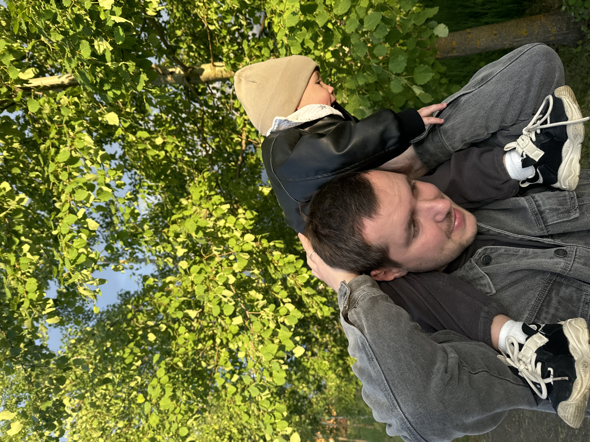 A man carries a child on his shoulders, outdoors. The man, wearing a grey denim jacket over a dark shirt, is facing towards the right of the frame and slightly upwards. The child, dressed in a beige beanie and a black jacket, is looking upwards and to the left. The background shows green foliage, possibly trees, with glimpses of blue sky. Based on the available information, the location is Vysokinichi, Russia.