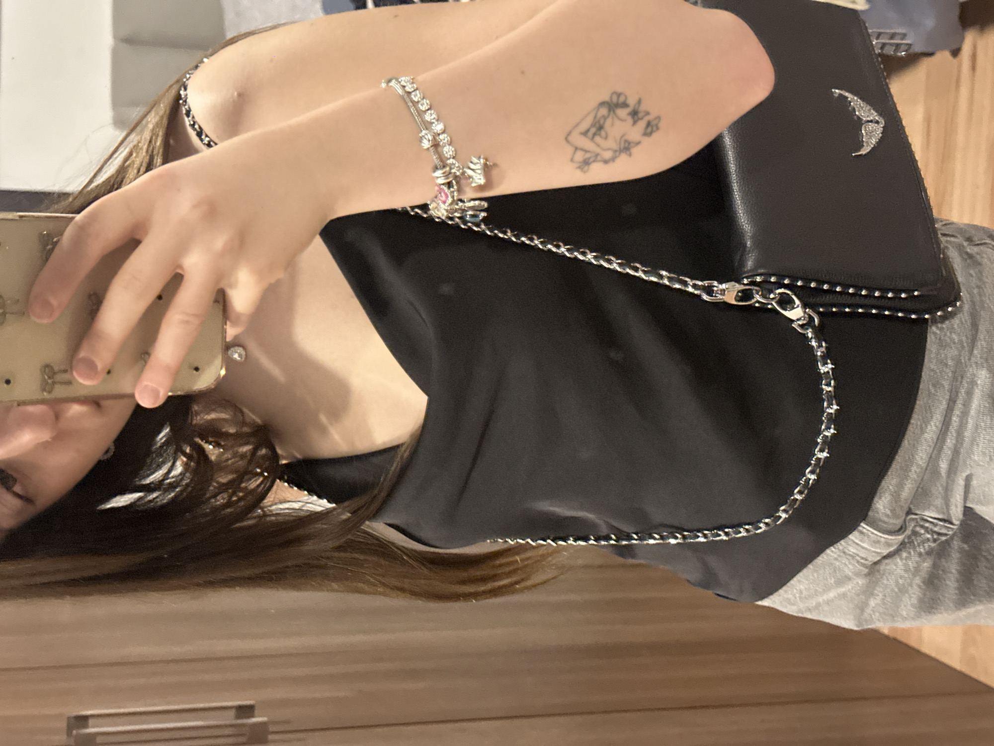 A person is taking a selfie indoors, holding a beige cell phone. The subject has shoulder-length brown hair and is wearing a black top. Visible on their left arm are a charm bracelet and a tattoo. A black purse with a silver chain is draped across their body. The location appears to be a room with a wooden floor and light-colored walls. There is no indication of Huixquilucan de Degollado, Mexico.