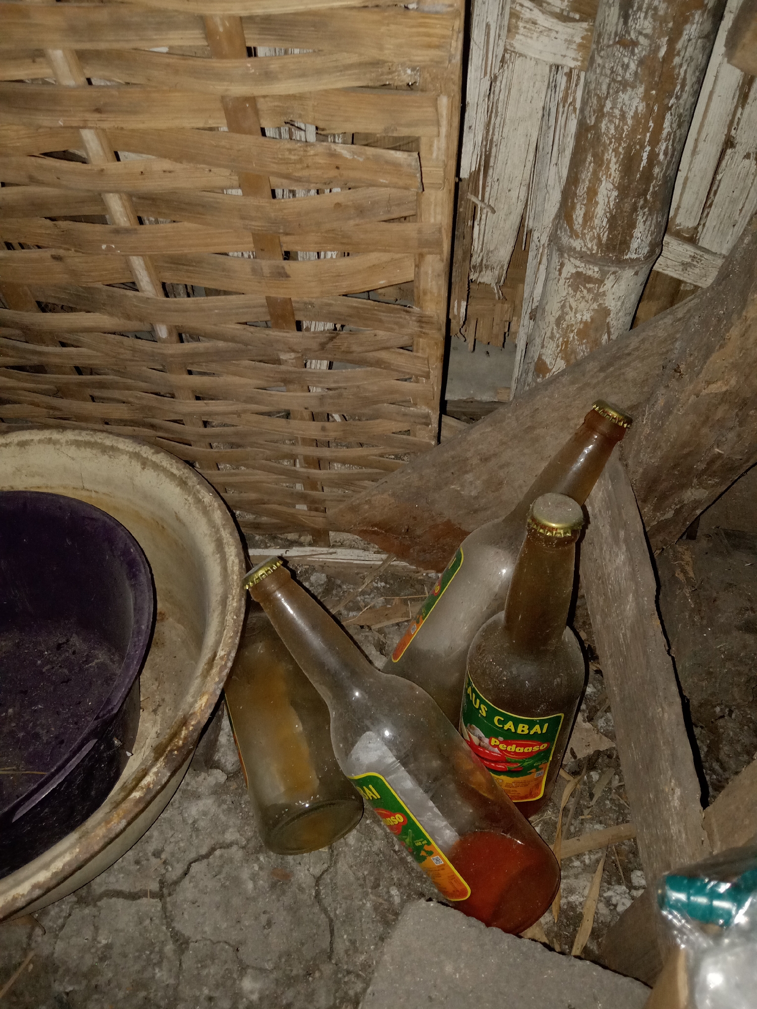 A close-up, low-angle shot captures a collection of discarded glass bottles and a plastic basin on a cracked concrete floor, leaning against a rustic woven bamboo wall and wooden beams. Three brown glass bottles are prominently featured. One bottle, upright and in focus, displays a label that reads "AUS CABAI Pedaaaso." This bottle appears to contain a reddish liquid. Another bottle, slightly out of focus, leans to the left, its contents less clear. A third, mostly empty, amber bottle lies on its side in the foreground. To the left, a large, worn, cream-colored basin holds a dark purple object, possibly another basin or bowl, within it. The background consists of a textured woven bamboo structure and rough wooden planks, suggesting an outdoor or semi-outdoor storage area. The lighting is dim, creating shadows that enhance the gritty, neglected atmosphere. The scene is consistent with a back alley or storage shed in a residential area of Jumput, Indonesia.