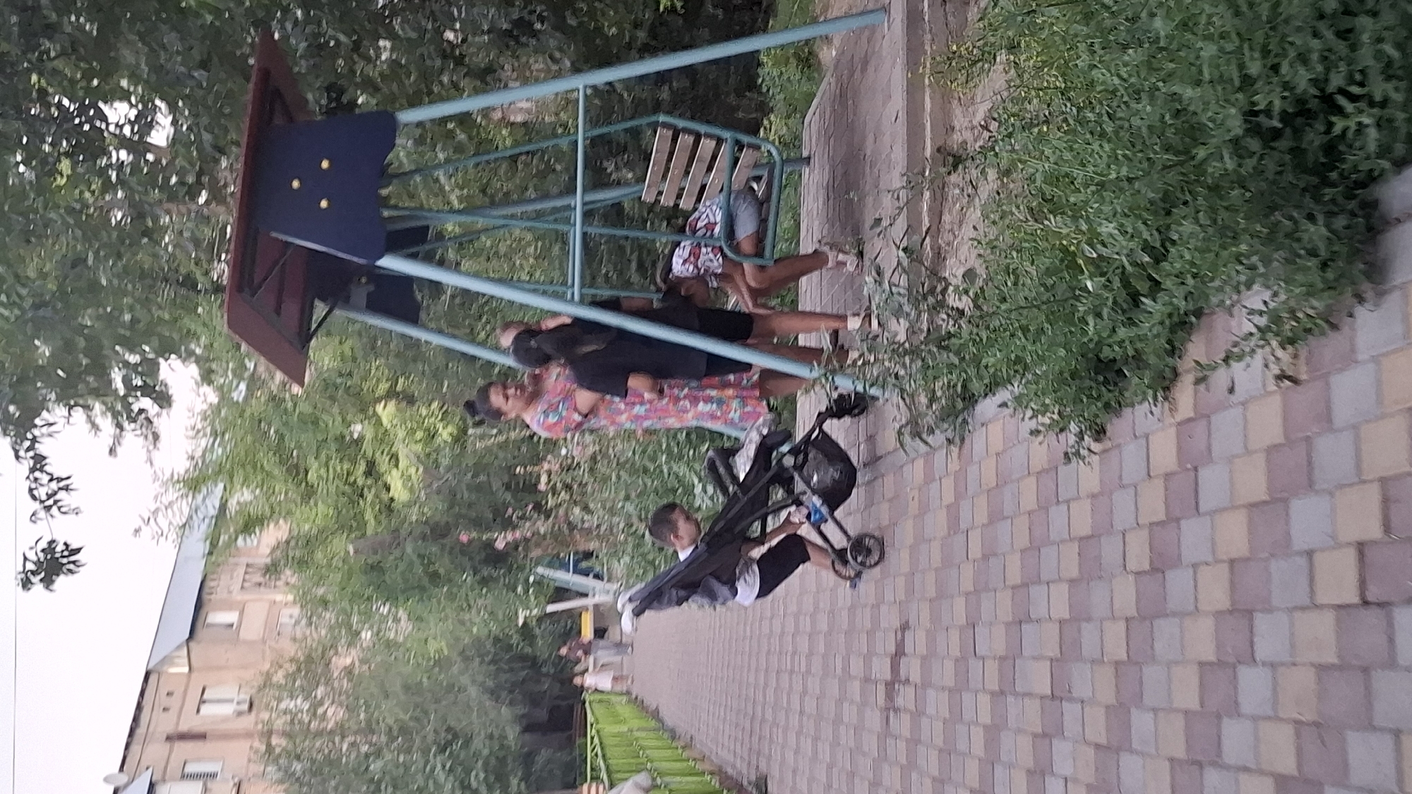 An outdoor scene depicts a group of people near a playground structure with a swing set. In the foreground, a young boy in a white t-shirt and black shorts is interacting with a stroller on a paved pathway. Behind him, a woman wearing a colorful patterned dress stands with two children. One child, a girl with dark hair pulled back, is wearing a black t-shirt and shorts, and another child, a girl with dark hair, is seated on the swing, leaning forward with her head down. She is wearing a white and black patterned shirt and denim shorts. The background shows a multi-story building and lush green foliage. The paved pathway is made of interlocking bricks in shades of red, beige, and grey. The playground has a blue and maroon swing set structure with a wooden bench. The scene appears to be in a residential area.