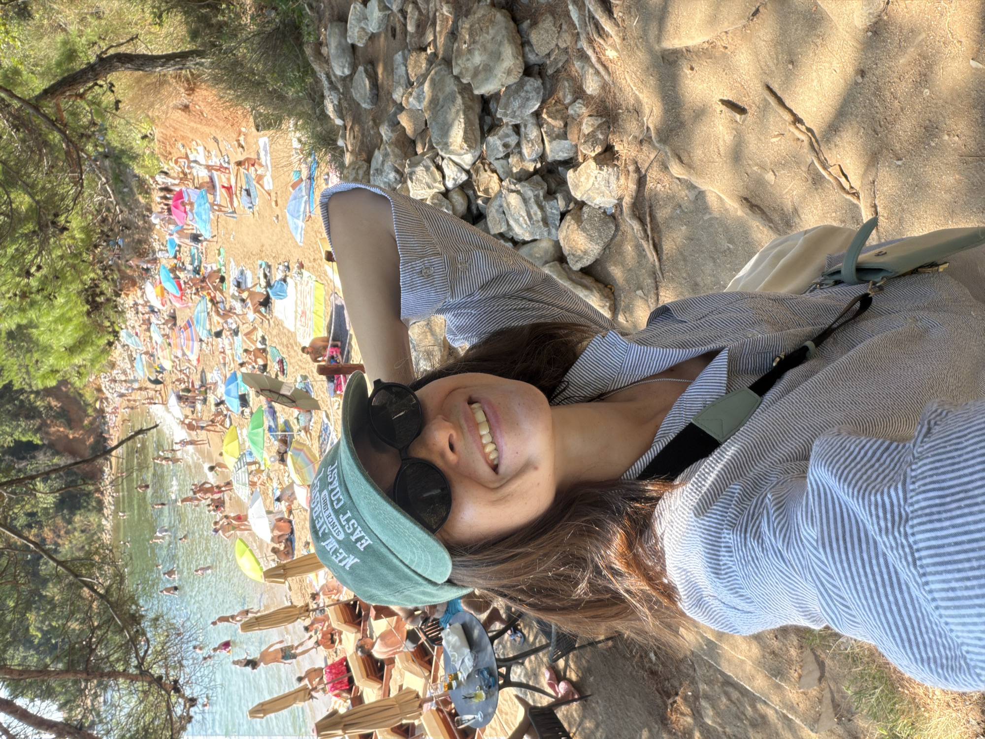 A medium shot captures a smiling woman wearing a green baseball cap and sunglasses, taking a selfie on a sunny day at a crowded beach. The woman has brown hair and is wearing a striped button-down shirt. In the background, the beach is filled with people sunbathing on lounge chairs, swimming in the clear blue water, and relaxing under colorful umbrellas. The terrain behind the woman is rocky and covered with dry roots, typical of a Mediterranean coastline. The scene appears to be located in Budva, Montenegro, a popular tourist destination known for its beautiful beaches.