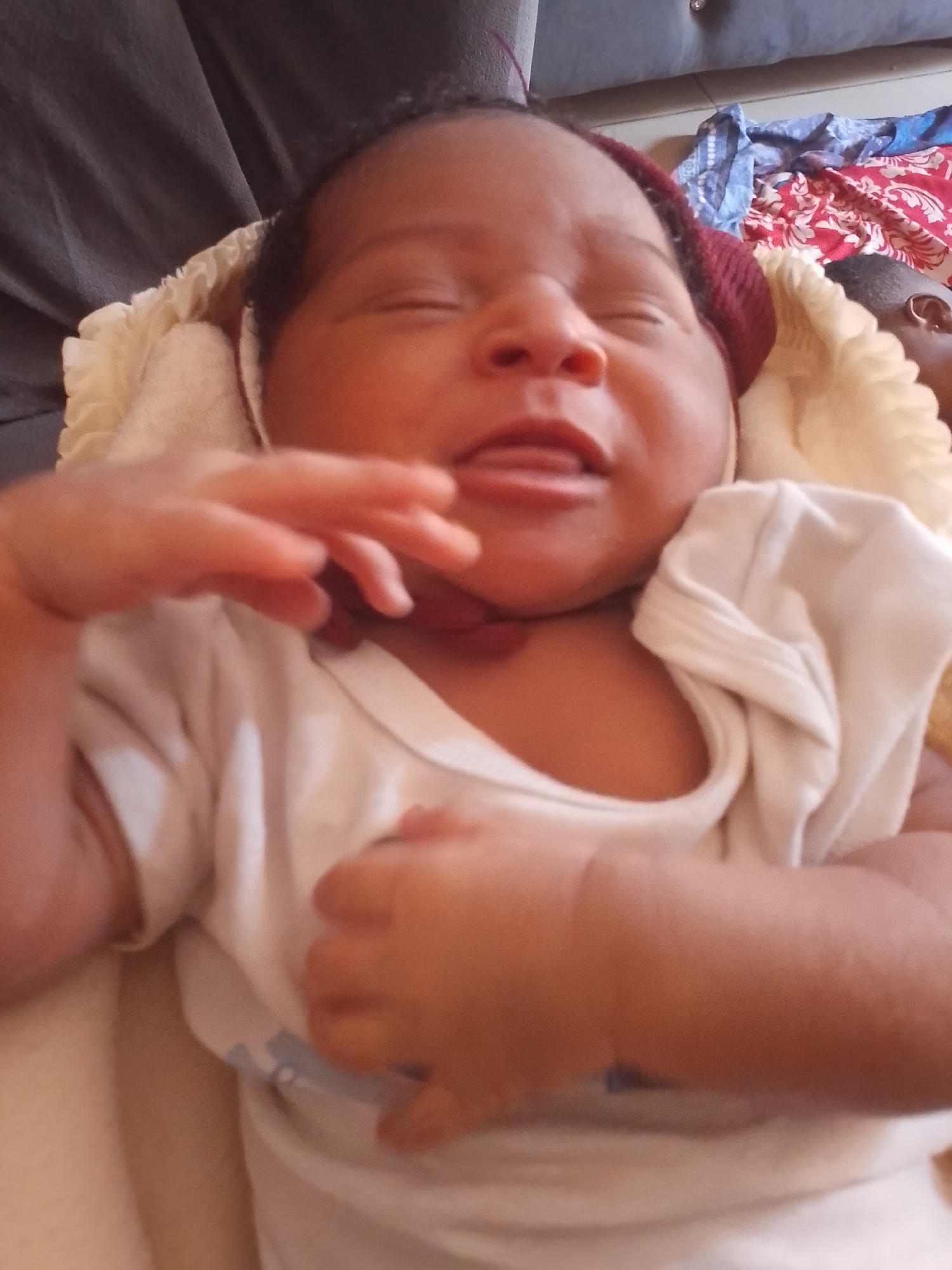 A newborn baby is lying on a soft, light-colored blanket. The baby is wearing a white shirt and a dark-colored knitted cap. The baby's eyes are closed, and their tongue is slightly protruding. The baby's hands are positioned near their chest, with fingers slightly curled. The background includes a dark-colored piece of furniture and some folded colorful fabrics. The location is Unknown, Unknown.