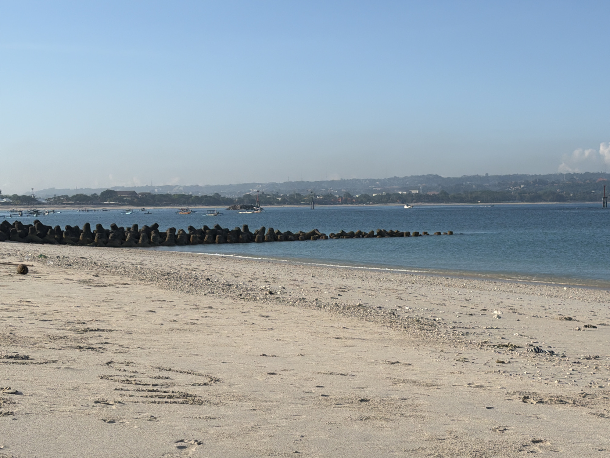 The image depicts a beach scene in Kuta, Indonesia. The foreground consists of a sandy beach with visible traces of waves and sand patterns. A line of tetrapod breakwaters extends into the water, protecting the shoreline. In the distance, the coastline shows a silhouette of land with buildings and vegetation. The water is calm, with a few boats visible. The sky is clear with a slight haze. A coconut sits on the shore.