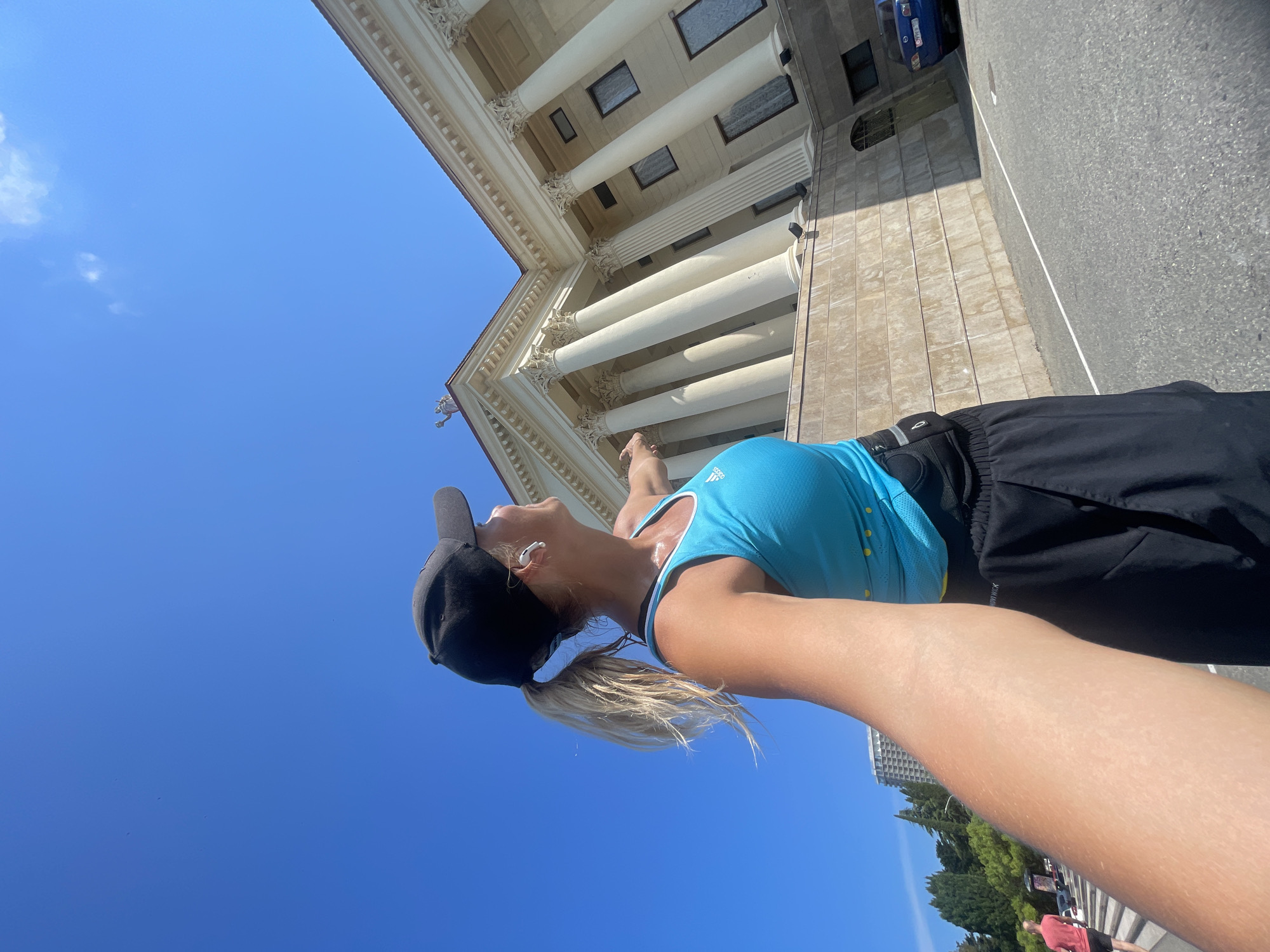 A woman wearing a black baseball cap, a blue athletic tank top, and black athletic shorts is pictured from a first-person perspective, as if taking a selfie while jogging. She is outdoors on a sunny day with a clear blue sky. She is looking to her right and pointing towards a large building with white columns and classical architecture. The building appears to be made of stone and has several windows. In the background, there are trees, other buildings, and a blue car parked on a road. The woman has blonde hair in a ponytail and is wearing white earbuds. The scene is set in the city of Sochi, Russia, specifically in front of the Dendrarium (The Arboretum) building.