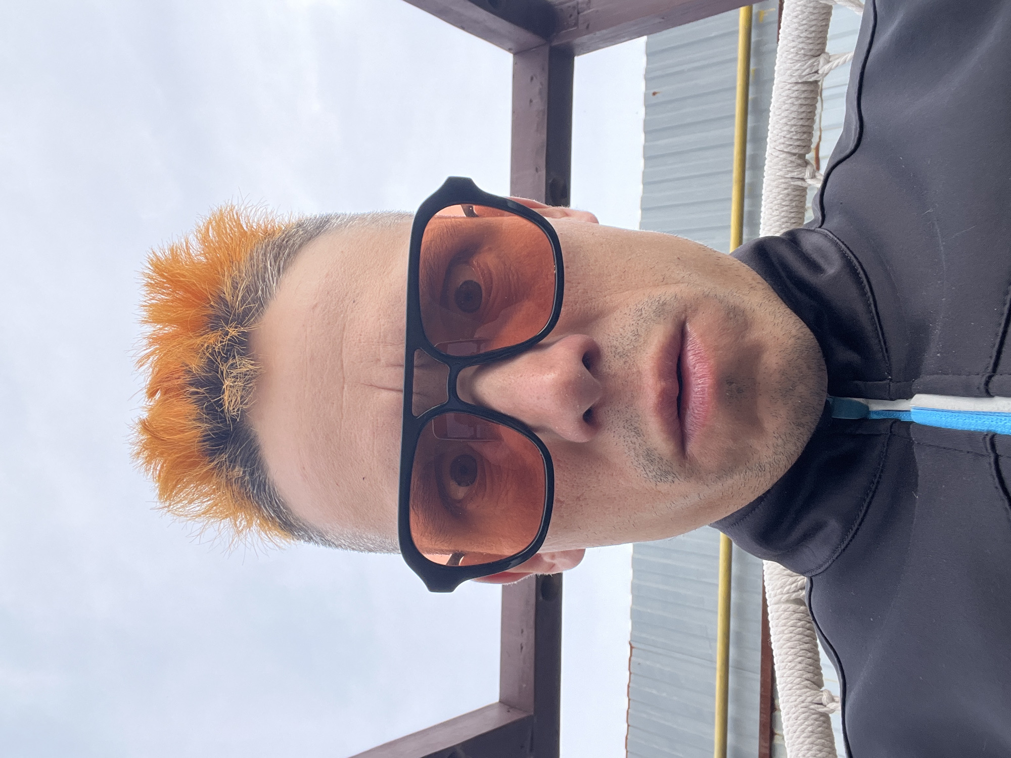 A man wearing black sunglasses and a black jacket with a blue and white stripe is looking directly at the camera. His hair is short and dyed orange. He appears to be outside, possibly in an area with some industrial-looking elements such as corrugated metal siding and wooden beams. A thick, woven rope is visible behind him. The location is Chaikovsky, Russia.