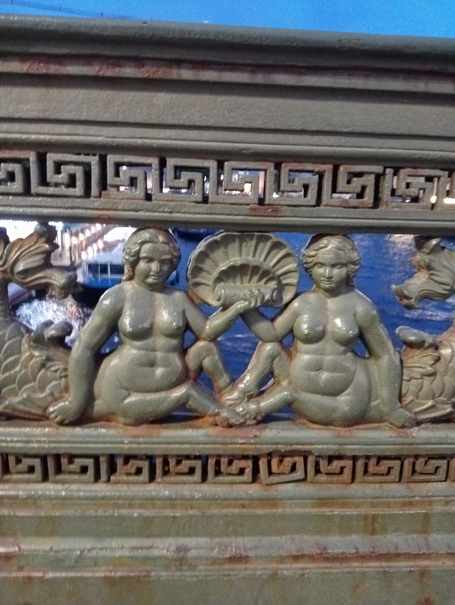 The image shows a section of a decorative metal railing, likely from a bridge or embankment in Saint Petersburg, Russia. The railing features a Greek key or meander pattern along the top and bottom edges of the central section. In the center, two nude figures, resembling mermaids or nymphs, are depicted. They are seated with their legs bent and are holding fan-like objects. The figures are detailed with rounded forms. To the left and right of the figures are stylized dragon or sea monster heads. The background appears to be water, possibly a canal or river, with hints of reflected lights from buildings on the opposite bank, suggesting an urban setting at night or dusk. The metal itself shows signs of weathering and rust, indicating age and exposure to the elements. The overall impression is of an artistic and historical embellishment to a piece of urban infrastructure.