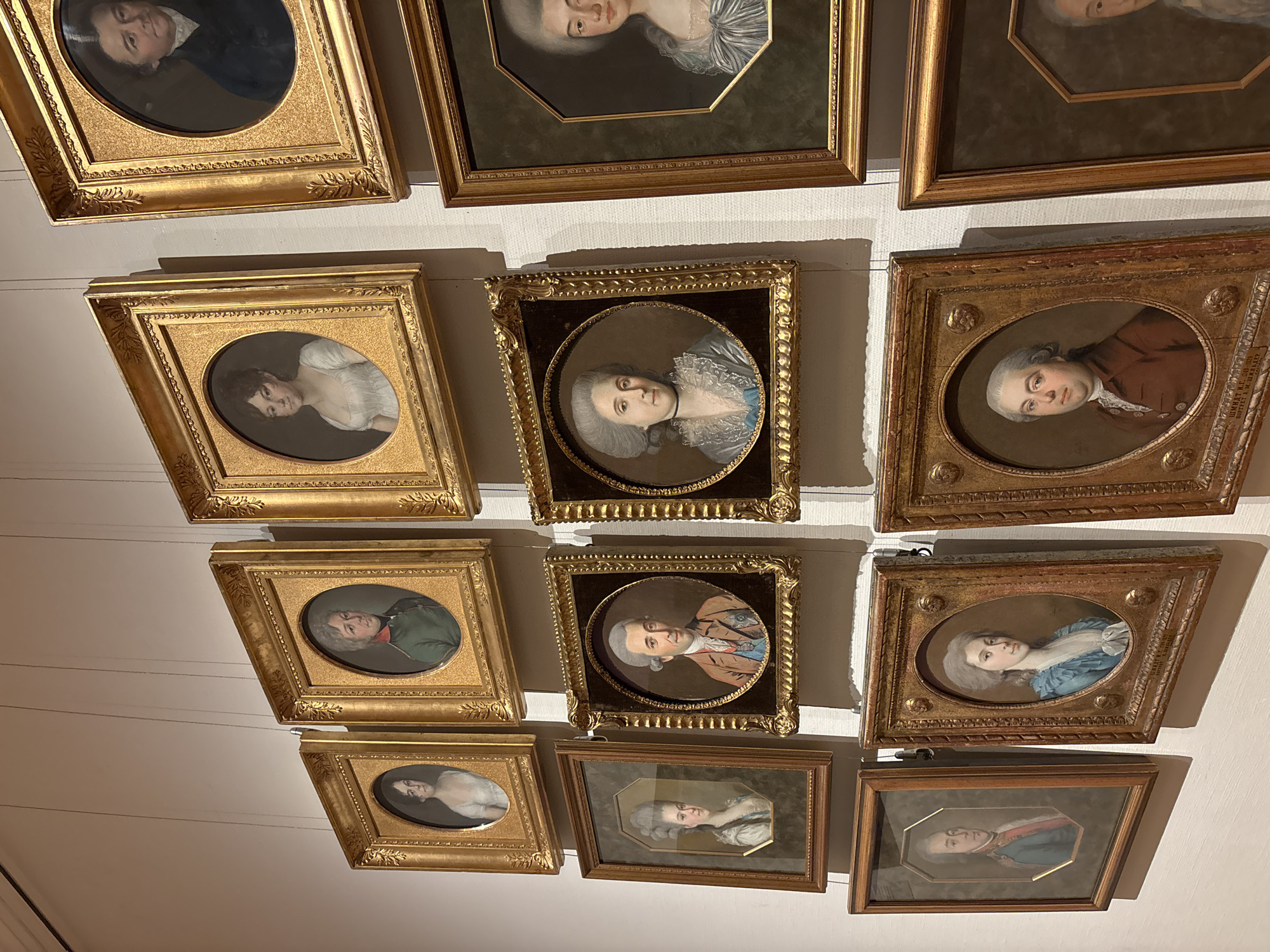 The image shows a collection of framed portrait paintings displayed on a white wall. The portraits appear to be of individuals from the 18th or 19th century, based on their clothing and hairstyles. The frames are mostly ornate and gold-colored, but some have a simpler, wooden finish. The portraits themselves are painted in a realistic style, likely pastel or watercolor miniatures, and depict subjects of various ages and social classes. The paintings are arranged in a grid-like pattern, with some overlap. There is a subtle indication of hanging wires, suggesting the pictures are actually displayed. The location is likely a museum or gallery in Moscow, Russia, displaying historical portraits.