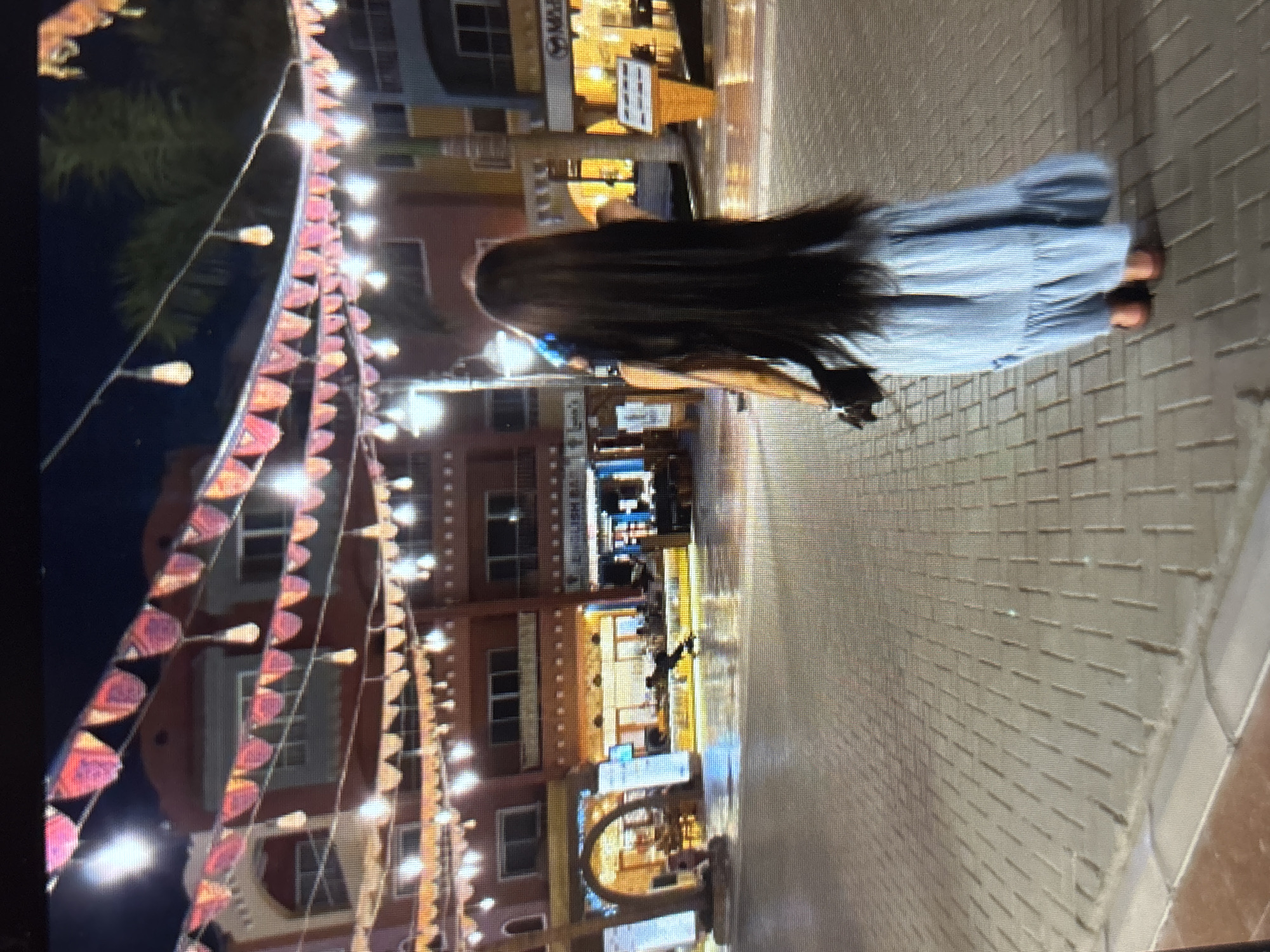 An outdoor nighttime shot shows a woman from behind, walking on a paved area in a city. She has very long, dark hair and is wearing a light blue striped dress with ruffles at the hem and flat, brown shoes. She is carrying a black clutch bag in her right hand. In the background, buildings are lit up and adorned with strings of small red flags and light bulbs. The pavement is wet, reflecting the lights. In the distance, a person is visible on a skateboard.