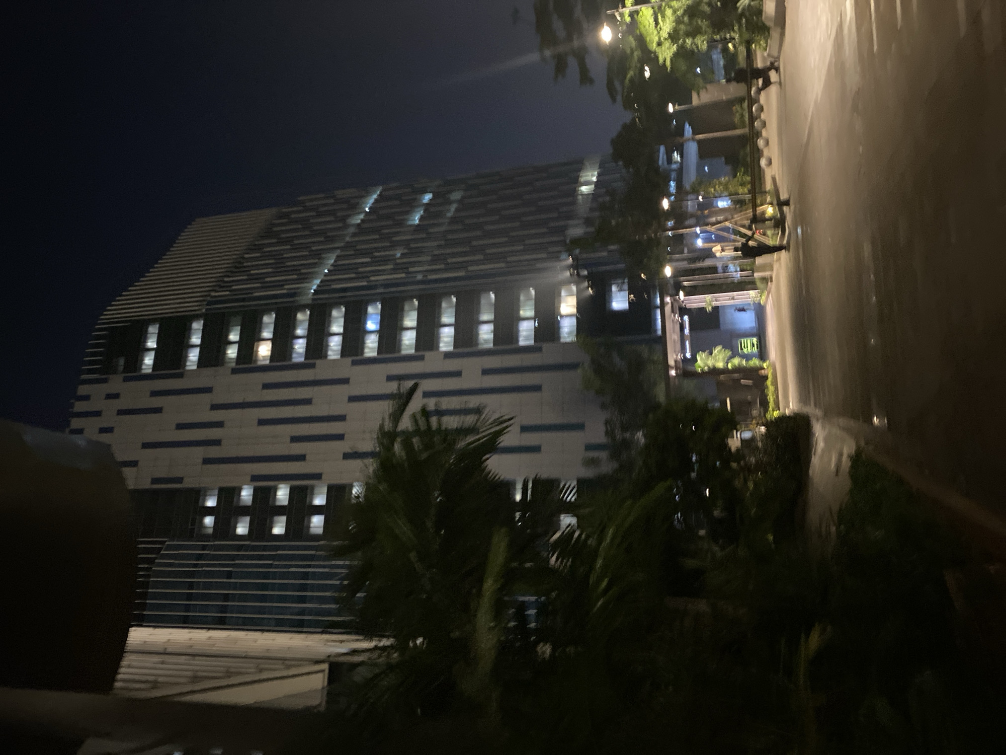 The image shows a modern skyscraper at night, likely in Central Jakarta, Indonesia. The building has a distinctive facade with vertical blue and white elements. Its many windows are illuminated, suggesting it is occupied. In the foreground, palm trees and other vegetation are visible, and a paved walkway or road leads towards the building. Streetlights and lights from within the building illuminate the scene. Two figures, appearing to be men in dark clothing, are present in the middle ground, one near a bench and the other by a barrier. The overall atmosphere is urban and contemporary, bathed in the artificial light of the night.