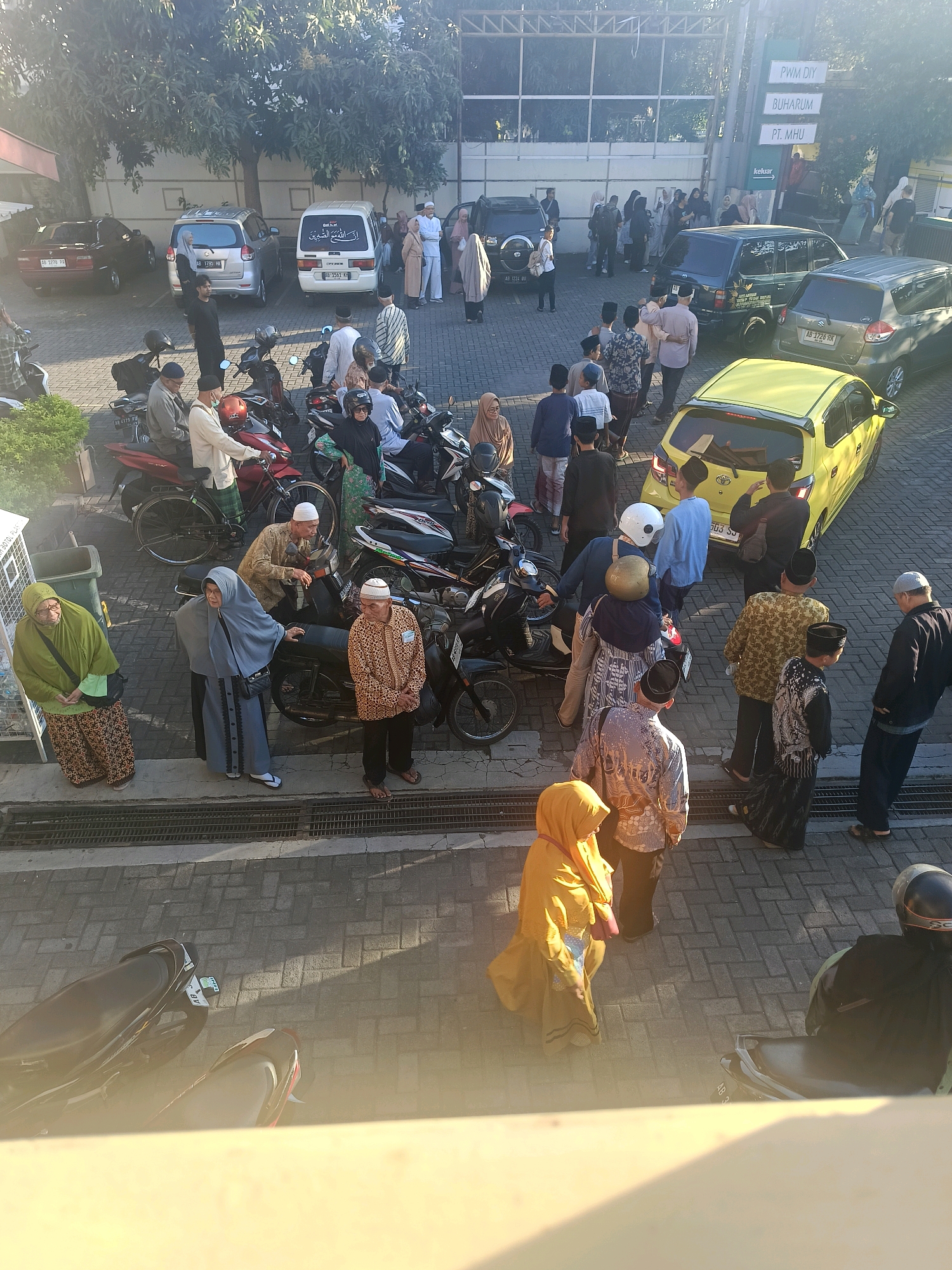 This aerial view captures a gathering of people, likely for an event or religious service, outside a building in Yogyakarta, Indonesia. The sun casts long shadows across the brick-paved ground, illuminating several parked motorcycles and cars, including a yellow hatchback. Several individuals are dressed in traditional Islamic attire, with men wearing peci caps and patterned shirts, and women in hijabs. Some are engaged in conversation, while others appear to be waiting or observing. A sign on the building reads "PWM DIY BUHARUM PT. MHU", indicating the presence of local organizations or businesses. The environment suggests a public space, possibly a parking lot or courtyard, adjacent to a building with a covered walkway.
