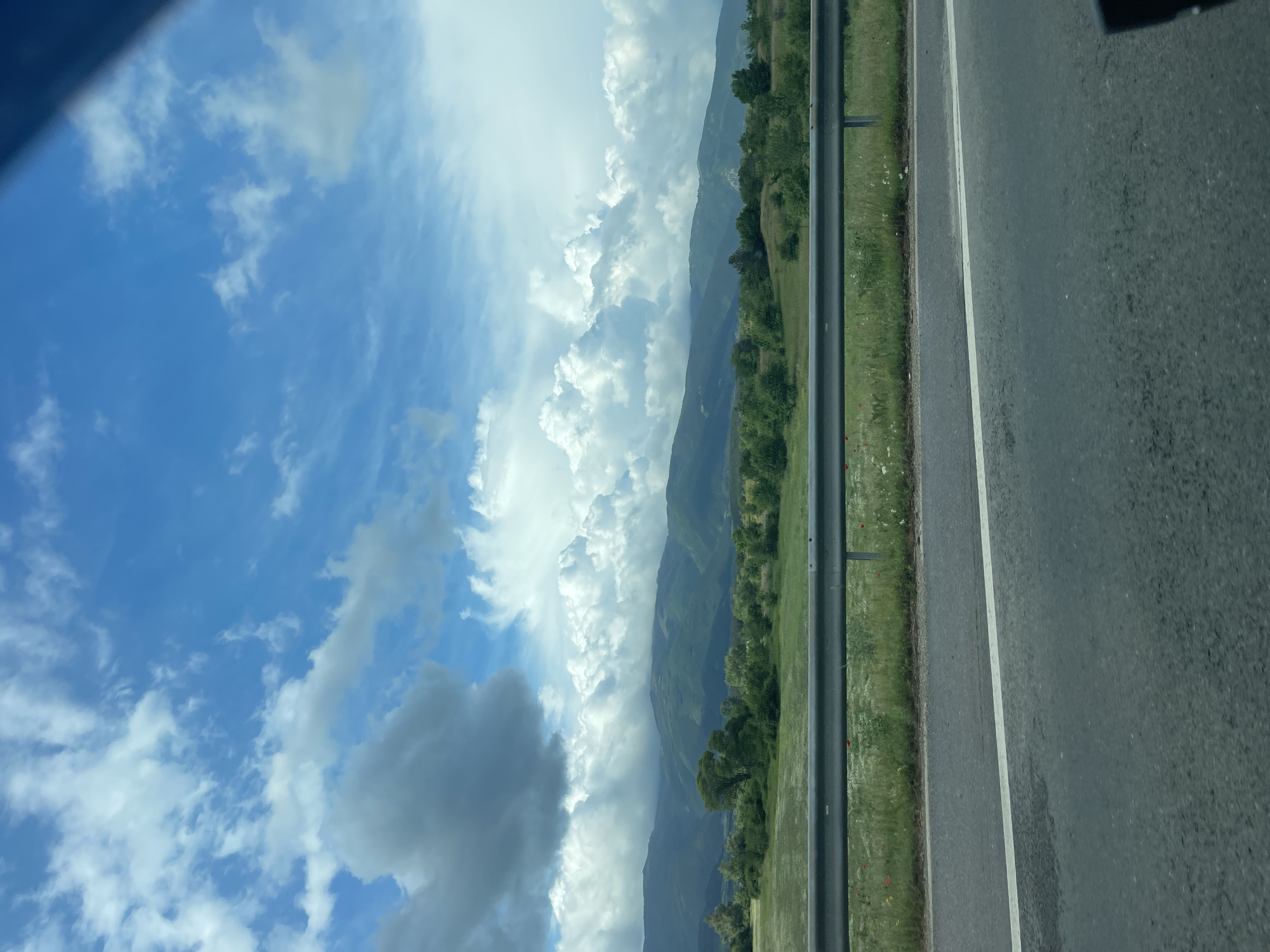 The image depicts a scene from a roadside. A segment of asphalt road, marked with a white line, occupies the right side of the frame. Adjacent to the road is a grassy shoulder. A metal guardrail runs parallel to the road and separates the roadside from a landscape of rolling hills and trees. Beyond the hills, the sky is filled with puffy white clouds against a backdrop of blue. The image does not show any people or any interactions and is taken in Konare, Bulgaria.