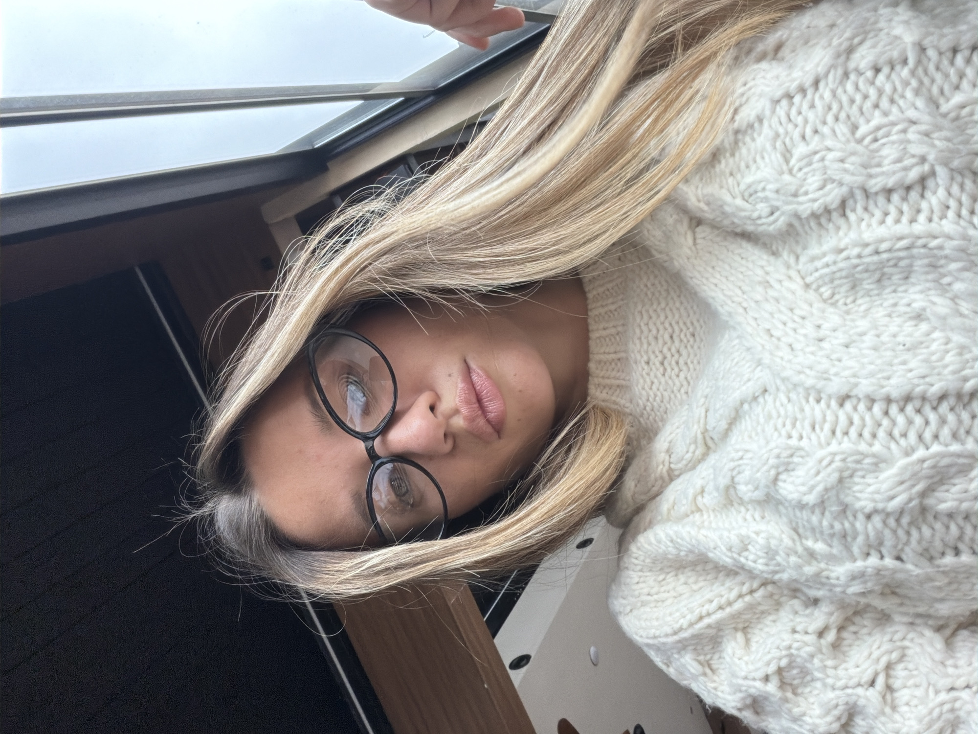 A female subject is captured in an upward-angled close-up, appearing to be a self-portrait. She has long, straight, light-brown to blonde hair with visible darker root growth and sporadic gray hairs near the crown. She wears black, oval-framed eyeglasses. Her lips are full and slightly parted, with a neutral expression. The subject is dressed in a thick, cream-colored cable-knit sweater with prominent patterns. The background indicates an indoor setting within the city of Unknown, Unknown. To the upper left, a dark ceiling with recessed lighting fixtures is visible. To the upper right, a large window with dark framing reveals an overcast sky. A light-brown wooden panel is present in the mid-background on the left. A partial hand is visible on the far right edge of the frame, near the window.