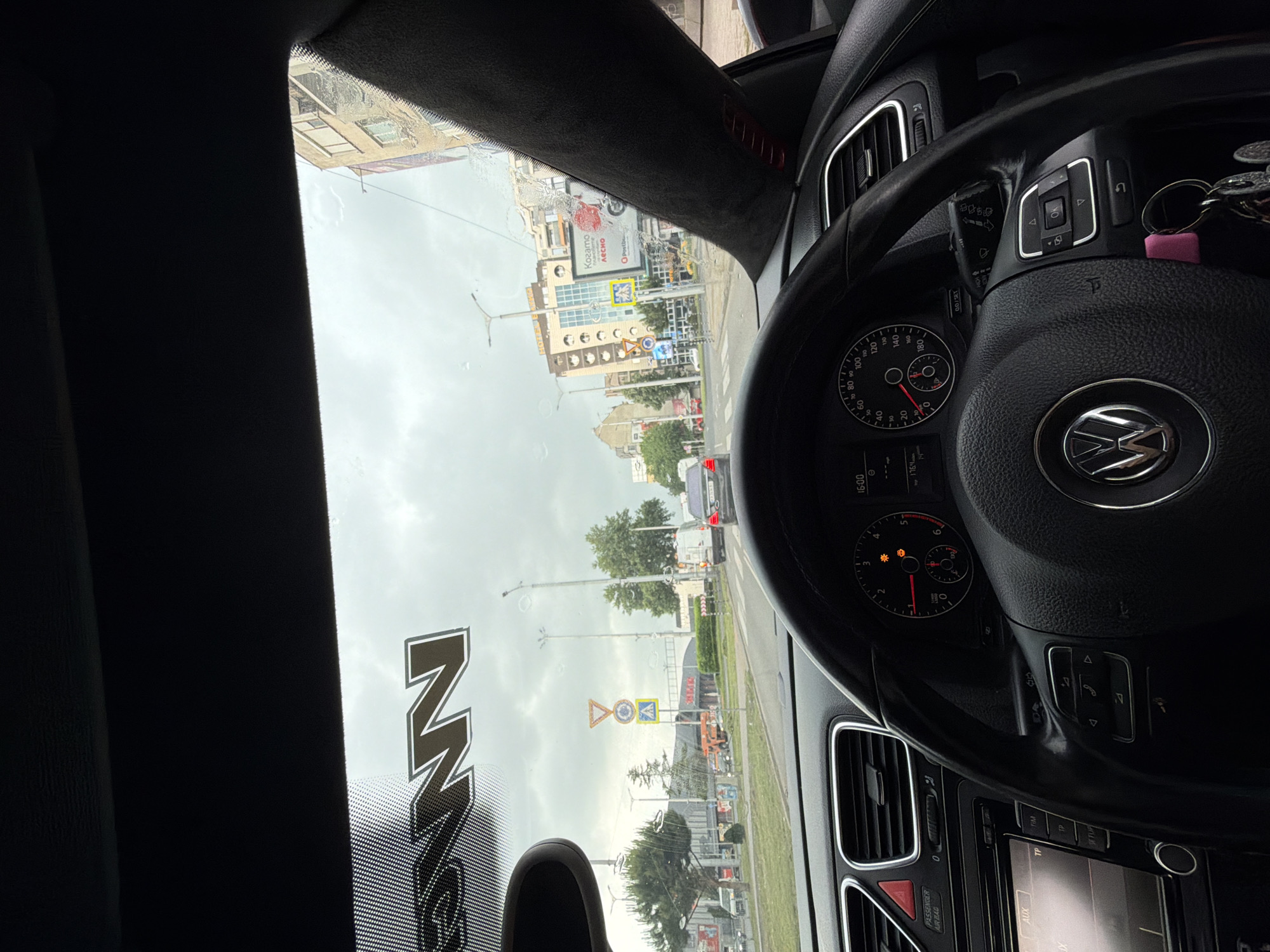 The image depicts a view from the interior of a car, looking out through a rain-splattered windshield onto an urban street scene in Plovdiv, Bulgaria. The perspective is slightly angled, suggesting the photo was taken by a passenger or from the driver's seat looking across.
Inside the car, a black leather steering wheel with a prominent Volkswagen logo is visible, along with parts of the dashboard and instrument cluster. The speedometer shows markings up to 200 km/h, and a digital display on the dashboard indicates the time as "16:00" (4:00 PM) and "1764", likely a trip meter or mileage. Keys with a pink keychain hang from the ignition. The infotainment system and air vents are visible below the steering wheel, with buttons labeled "AUX," "TIM," "TP," and "SETUP." The text "PASSENGER AIR BAG" is also present on the dashboard. A stylized "NN" or "MM" logo is visible as a dark graphic on the lower left portion of the windshield.
Outside, the weather appears overcast, and raindrops and smudges on the windshield suggest recent or ongoing rain. A city street with vehicles, streetlights, and buildings stretches into the distance. Notable structures include a multi-story building that partially shows "HOTEL..." and a large billboard. The billboard displays Cyrillic text, including "Когато" and "лесно," alongside "Postbank" and a logo for "ARCHO." Several road signs are visible, including a yellow diamond (priority road), a blue circular sign with a white arrow (mandatory direction), and a yellow square sign with a black pedestrian symbol (pedestrian crossing). The scene captures an ordinary afternoon in a bustling city environment.