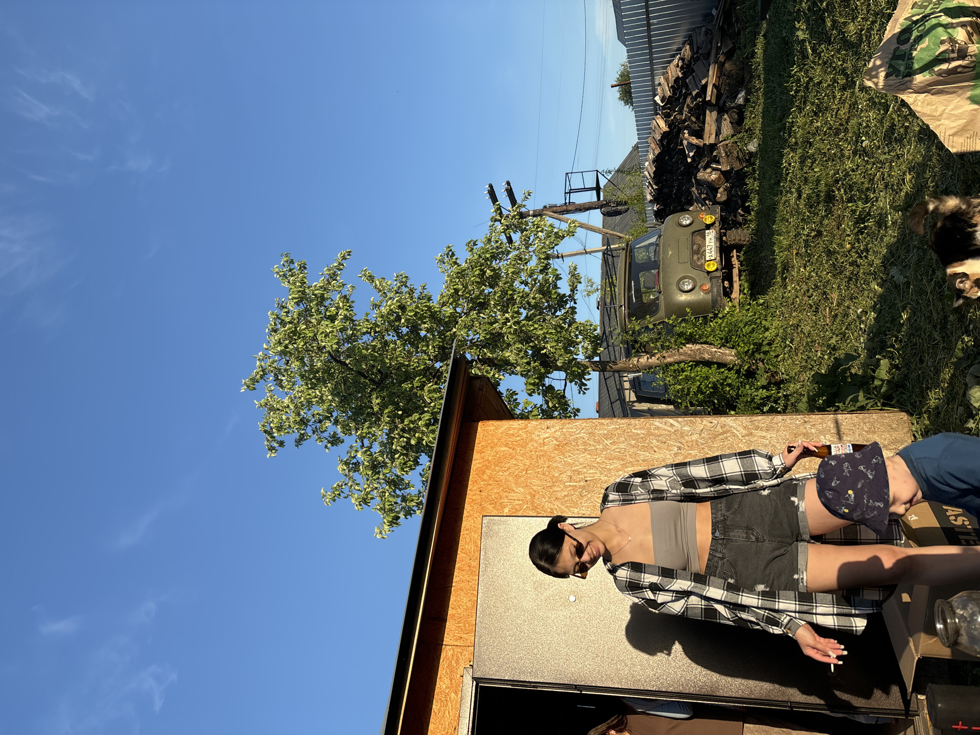 The image depicts a woman standing in front of a small, unfinished wooden structure with a metal door. She is wearing a grey tank top, black shorts, and a black and white plaid shirt. She is holding a beer bottle and appears to be smoking a cigarette. Another person, partially obscured, is wearing a blue shirt and a baseball cap. They are standing next to a cardboard box.
In the background, a green military vehicle (likely a UAZ-469 or similar) is parked near a pile of firewood, with overgrown grass and a chain-link fence visible beyond. Power lines stretch across the sky. The weather appears to be sunny, with a clear blue sky.
While the image is taken in an outdoor setting, there is not enough information to verify the location with certainty within the city of Фоки, Russia.