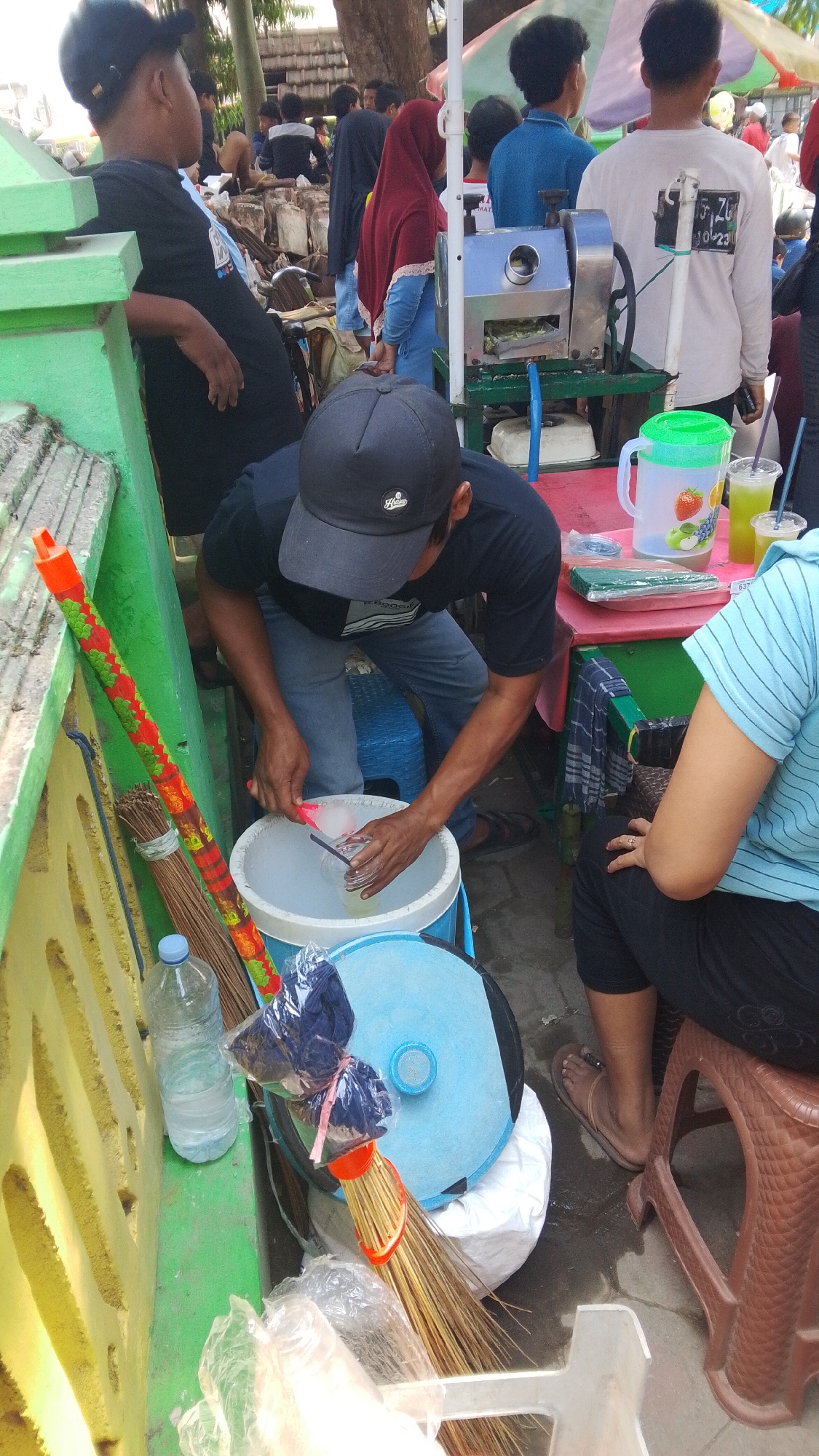 The image depicts a street scene in Mancar, Indonesia, likely a market or roadside stall. In the foreground, a man wearing a black cap and t-shirt is preparing drinks, scooping ice into a glass with a pink scoop. Beside him is a large white bucket filled with ice and water, a broom with a colorful handle, and a plastic bottle of water. A woman in a blue striped shirt and black leggings sits on a brown stool to the right, facing away from the camera.

Behind them, a stainless steel sugarcane juicer is visible, suggesting this is a beverage stall. Several other people are in the background, including a woman in a red hijab and a man in a blue shirt operating the juicer. The environment appears to be outdoors, with some greenery and what looks like a temporary structure or awning overhead. The ground is paved with concrete or similar material. The overall scene conveys a busy, informal commercial setting.