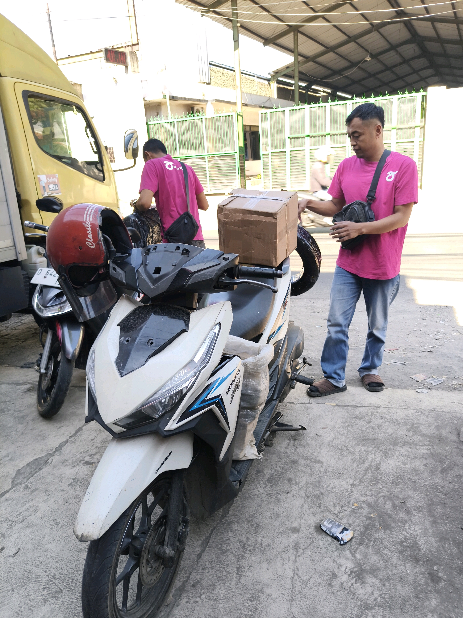 In Majasetra, Indonesia, two men are interacting with packages and motorcycles. One man, wearing a pink t-shirt and blue jeans, stands beside a white motorcycle, holding a brown cardboard box. He has a black sling bag across his chest. The other man, also in a pink t-shirt, is partially obscured by the first man and is near a yellow truck. A red helmet rests on the handlebars of a dark-colored motorcycle to the left. The scene appears to be outdoors, possibly in a delivery or logistics area, with a covered structure visible in the background and a green metal fence. The ground is paved with concrete.