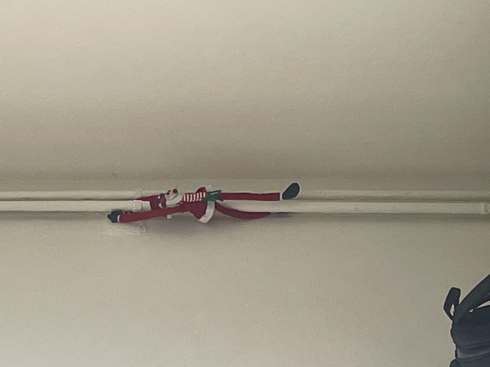 An Elf on the Shelf doll is lying horizontally across a white pipe attached to a beige wall. The doll has a red suit with white trim, black mittens and shoes, and a white face with a red nose. A striped scarf is around the doll's neck. Part of a black object is visible in the bottom right corner. The location in the city of Burgas, Bulgaria cannot be determined from the image.