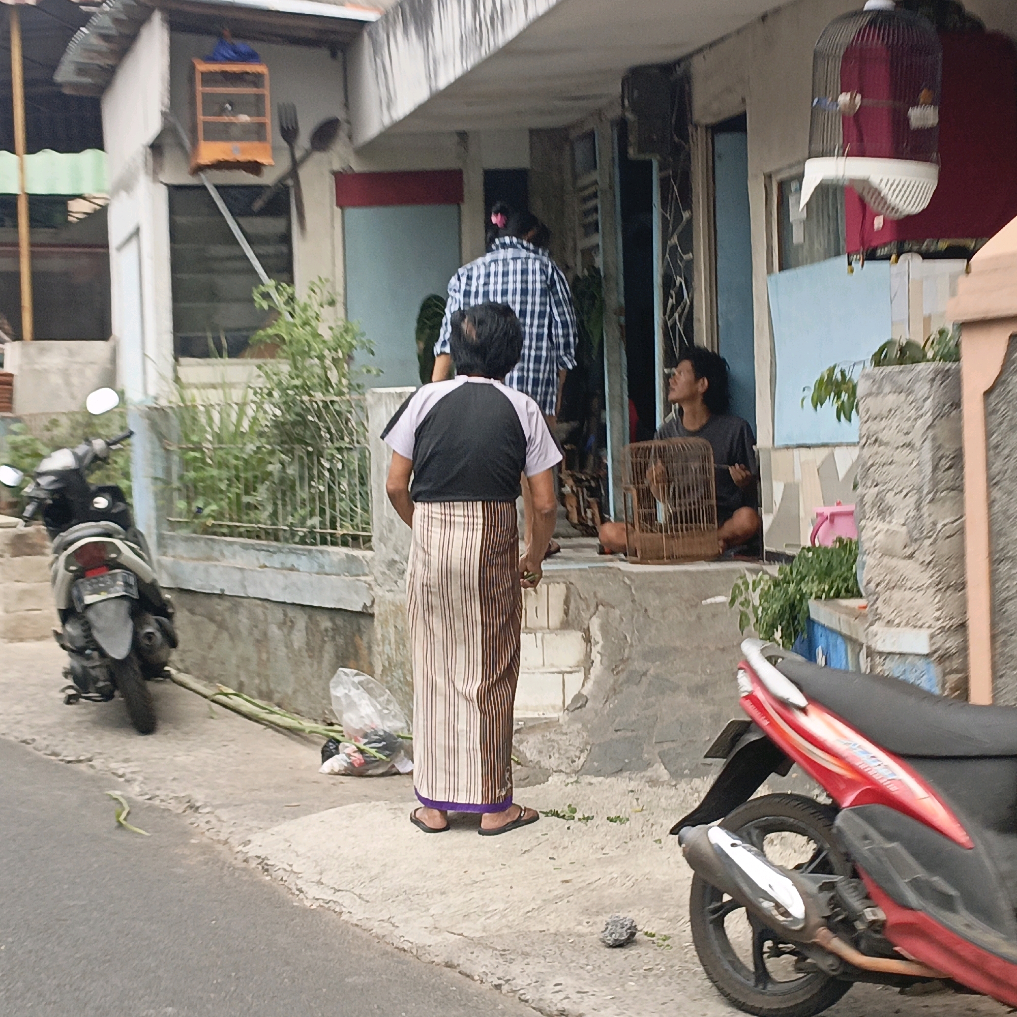 The image depicts an outdoor scene in what appears to be a residential or commercial area in Salatiga, Indonesia. In the foreground, a red and black scooter is partially visible on the right side of the frame, parked on a paved surface. To the left, another scooter, this one black and silver, is parked on the sidewalk.
A man wearing a black and white t-shirt and a striped sarong stands facing away from the camera, looking towards a building entrance. Behind him, a person in a blue and white checkered shirt is walking away. To the right, a man with dark hair is seated on the ground, looking upwards, next to a birdcage. A larger birdcage hangs from the building's overhang on the left.
The building appears to be two stories, with a concrete exterior and visible plumbing. There are plants and greenery around the building, and debris, including a plastic bag with flowers and a green snake-like object, is scattered on the ground. The overall impression is a casual, everyday street scene.