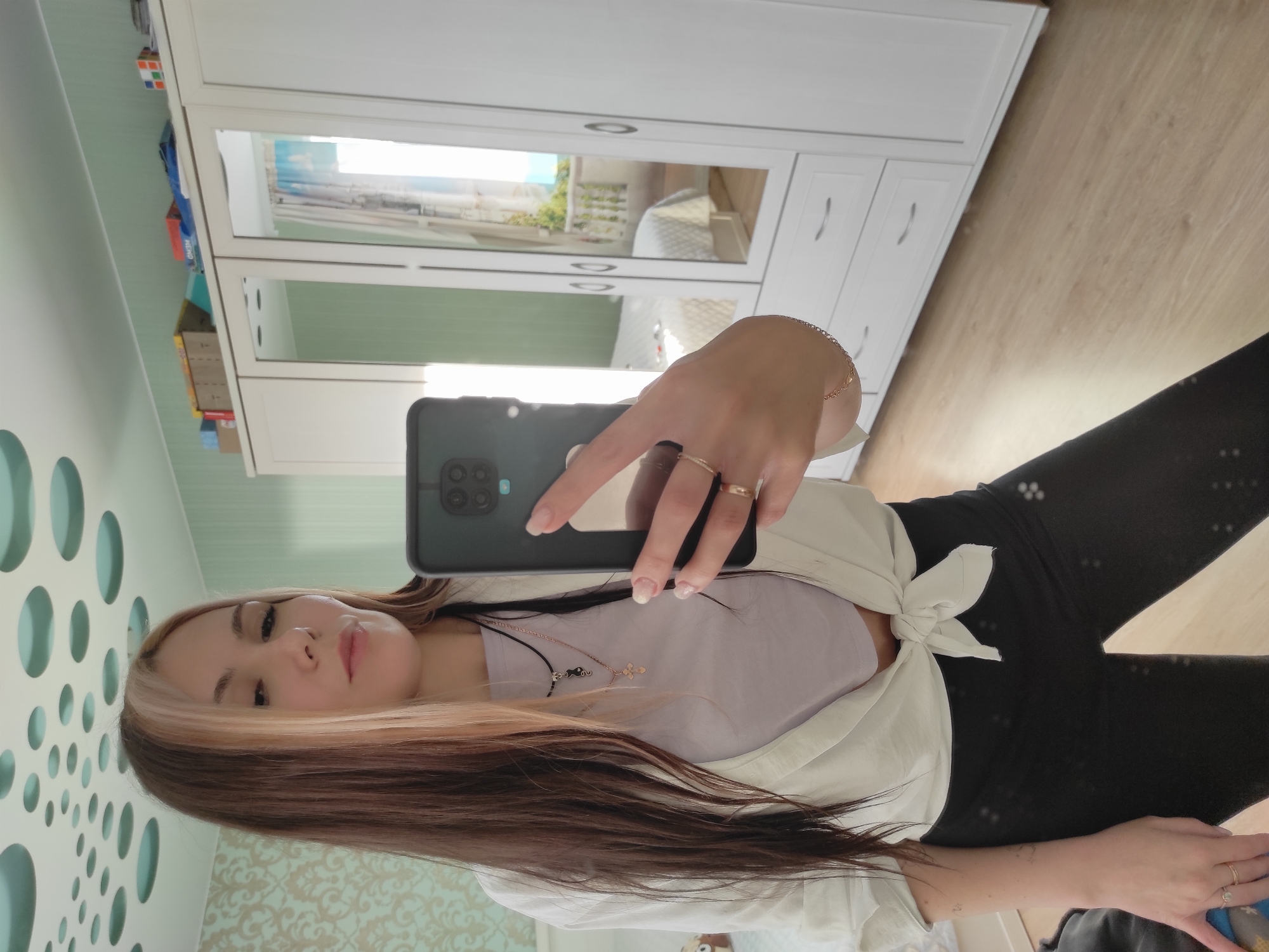 A selfie taken indoors in Ольховка, Russia, shows a woman with brown and blonde hair holding a black smartphone. She wears a light purple top and a white shirt tied in front. Rings are visible on her fingers, and two necklaces are around her neck. Behind her is a white wardrobe with mirrored doors, reflecting a bedroom interior. A patterned wallpaper is also visible. The floor appears to be wooden.