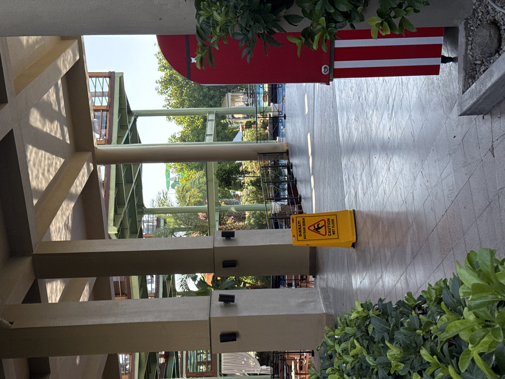 The image depicts an outdoor walkway at a resort in Aksu, Turkey. A yellow caution sign with the text "DIKKAT! KAYGAN ZEMIN" and "CAUTION WET FLOOR" is visible on the tiled ground. To the right of the sign is a red structure with vertical white stripes, possibly a trash receptacle or part of a decorative element. Lush green bushes line the left side of the walkway, and in the background, a swimming pool is visible along with palm trees and part of a resort building. The scene appears to be illuminated by natural daylight.