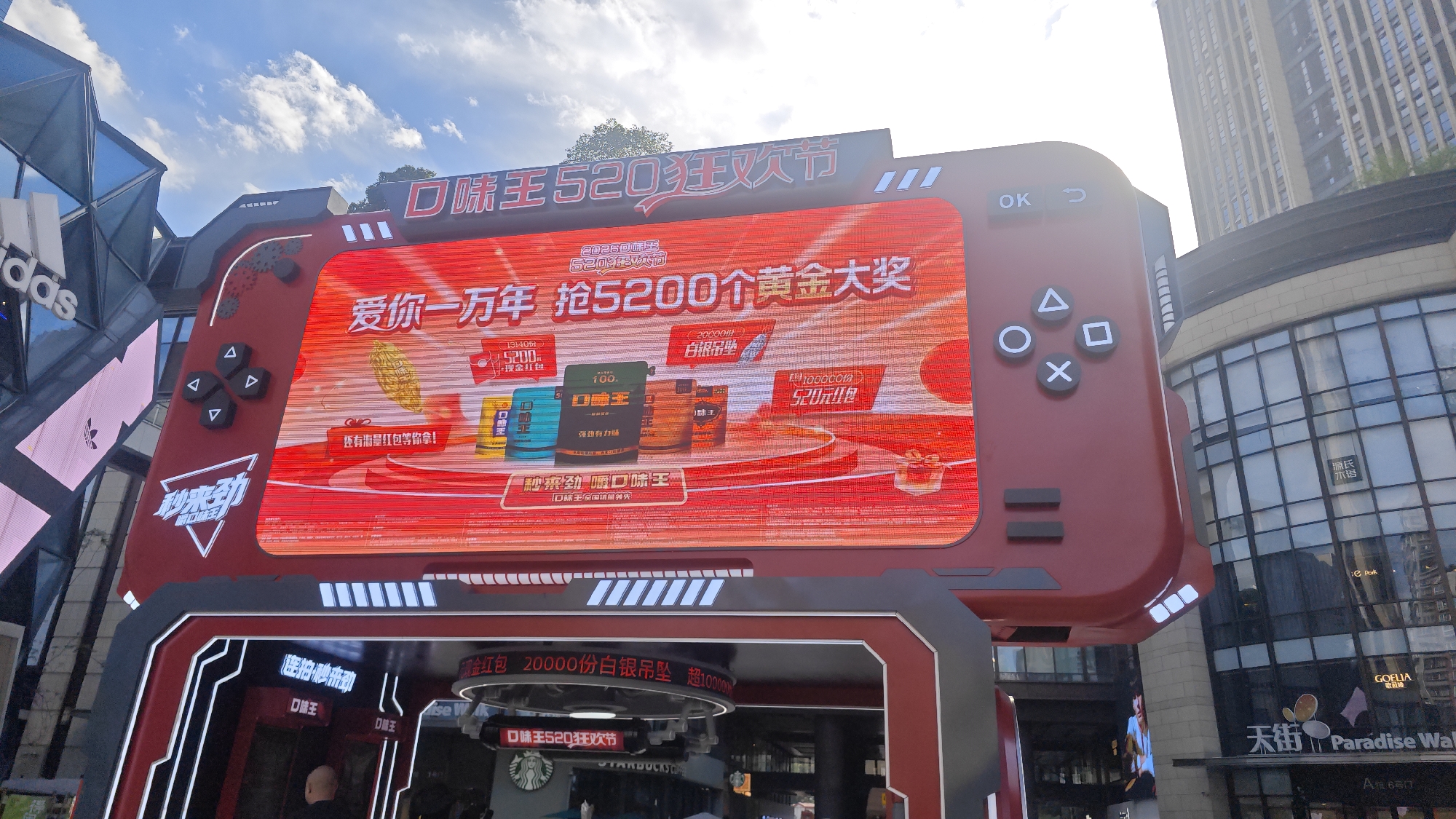The image depicts a large advertisement for "Kou Wei Wang" (口味王), a brand of betel nut, set up in a commercial area in Yuzhong District, China. The advertisement is designed to resemble a handheld gaming device, with a large screen displaying promotional information in Chinese, including phrases like "Love you for ten thousand years" and "Grab 5200 gold prizes." The screen also displays images of different packages of Kou Wei Wang products. The gaming device-shaped structure has buttons similar to those on a gaming console, such as a directional pad and buttons labeled "OK", triangle, circle, square, and cross.
Below the large screen is an entrance or storefront, seemingly affiliated with a Starbucks, with promotional text about Kou Wei Wang. The surrounding environment includes modern buildings, with retail establishments like Adidas and GOELIA visible. The area is identified as "Paradise Walk" or "Tianjie" (天街) in Chinese. There are people present, including a person entering the storefront below the advertisement. The sky is partly cloudy, suggesting it's daytime.