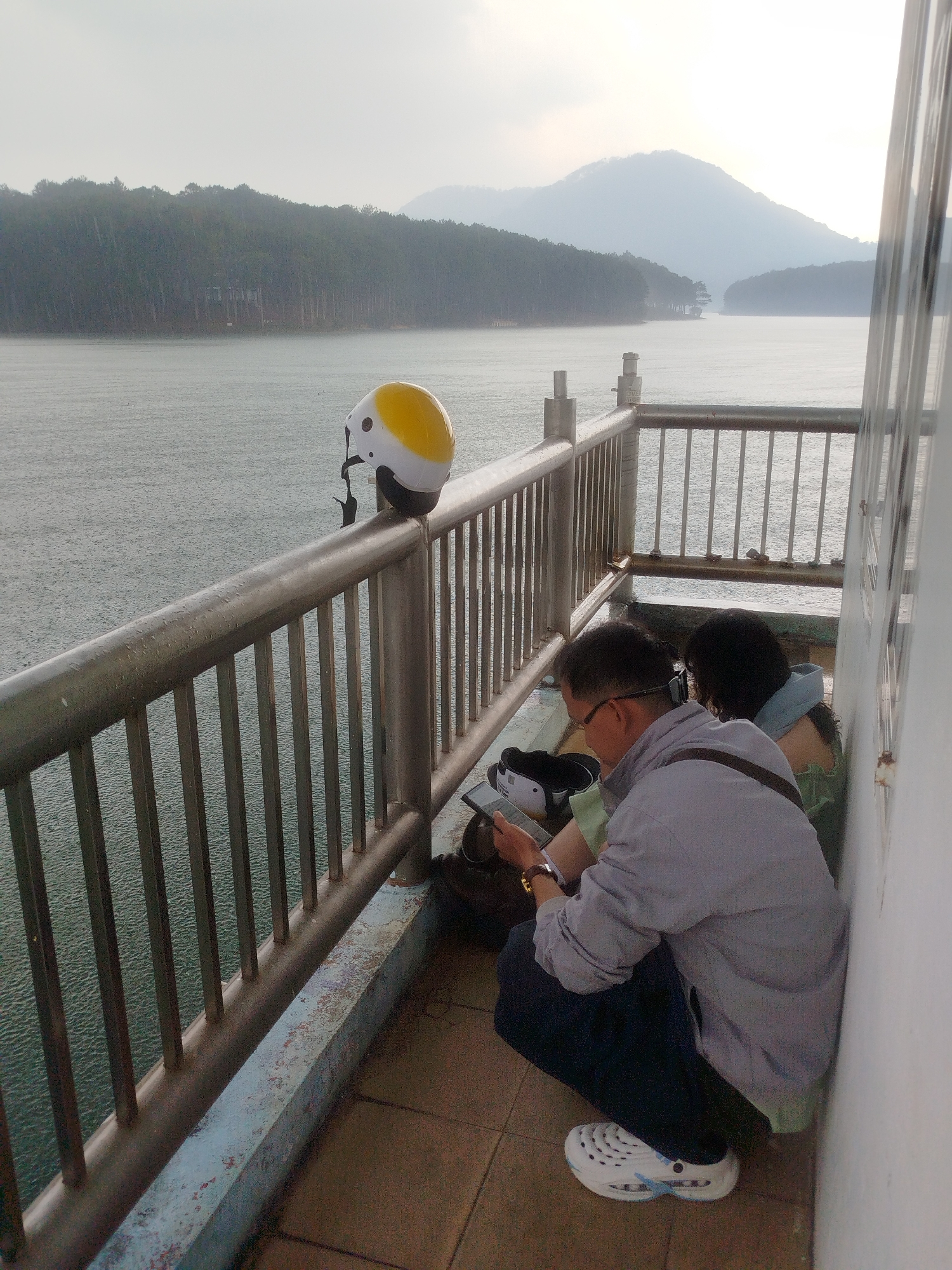 Two people are crouching on a tiled walkway next to a metal railing. One person, presumably male, is wearing glasses, a grey shirt, dark pants, and white Crocs shoes, and is looking at a mobile phone. The other person, possibly female, has dark hair and is wearing a green top and blue jacket. They are sitting against a white wall. A backpack, black shoes, and other items are on the ground near them. On the railing to their left is a yellow and white helmet. Beyond the railing is a large body of water under a cloudy sky, with forested hills in the distance. The location is likely Tuyen Lam Lake in Da Lat, Vietnam, given the scenery and weather. It appears to be raining or drizzling.