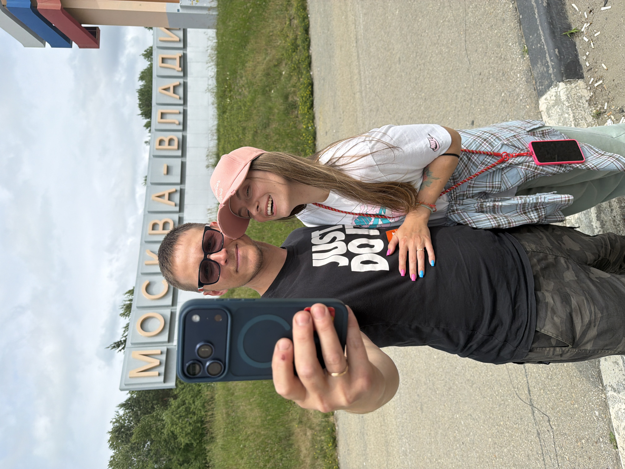 In a medium shot, a young couple takes a selfie in front of a sign that reads "MOSKVA-VLAD." The man, on the left, wears sunglasses and a black t-shirt with "JUST DO IT" printed on it, and camouflage pants. He is holding a dark blue smartphone, taking the photo. His left hand, which wears a gold wedding band, rests on the woman's shoulder. The woman, on the right, smiles as she leans into the man. She has a pink baseball cap on her head, and wears a white t-shirt with a graphic design, and a light blue plaid shirt tied around her waist. Her right hand, painted with bright pink and blue nails, is placed on the man's chest. A red cord hangs from her neck, attached to a pink phone case. The couple stands on a paved area next to a grassy slope. The sign is large and illuminated, with the letters outlined in gold. A tall, colorful monument stands to the right of the sign, partially visible in the frame. The sky is overcast. The location appears to be a roadside in Nikolaevka, Russia, indicated by the sign.
