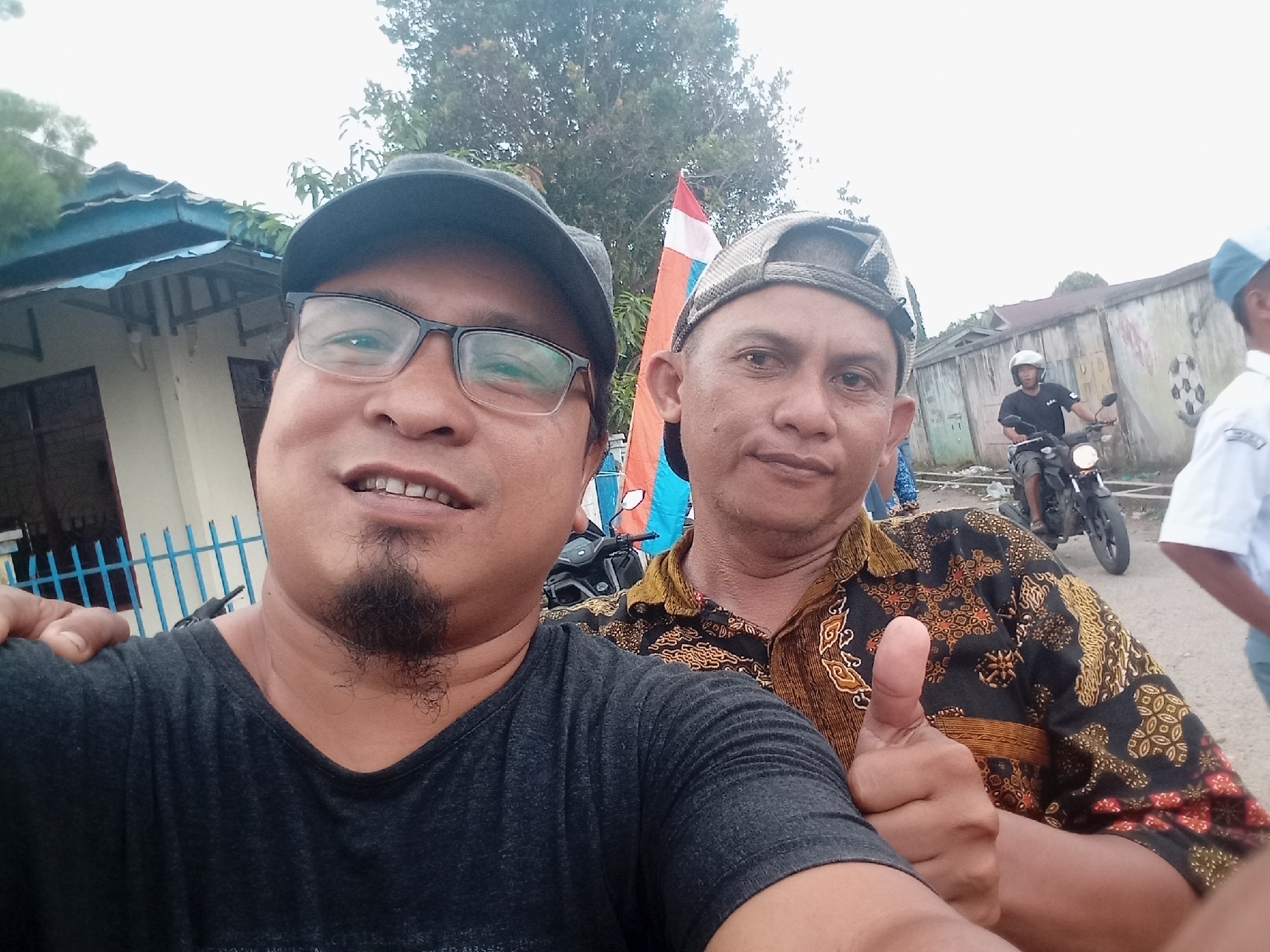 The image is a selfie taken outdoors in Candirejo, Indonesia. In the foreground, two men are visible. The man on the left, wearing glasses and a dark baseball cap, is in the process of taking the photo. He has a goatee and is wearing a dark t-shirt. The man to his right, wearing a patterned shirt and a baseball cap, is giving a thumbs-up. In the background, a red, white, and blue flag is visible. Further in the background, a person is riding a motorcycle. The scene appears to be a street with buildings and a fence.