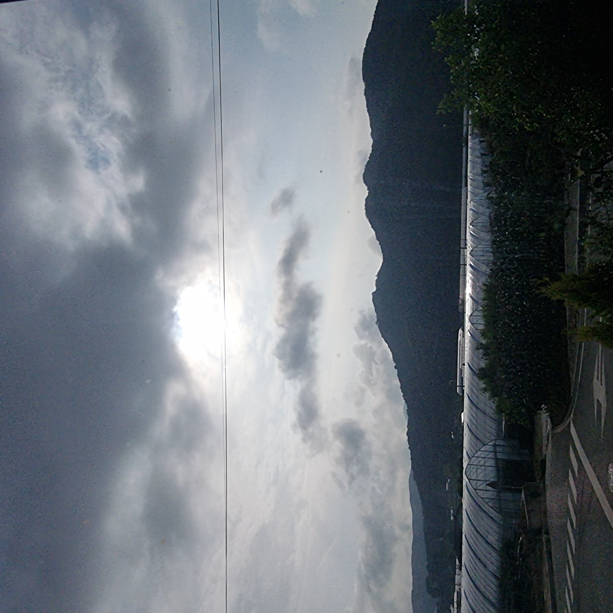 The image shows a cloudy sky with the sun peeking through in the upper center. In the middle ground, there is a range of dark, tree-covered mountains. In the foreground, there are several long, arched greenhouses with transparent coverings, stretching across the landscape. A road with white markings is visible in the lower left corner, and a dark green hedge or dense foliage borders the greenhouses. A train is visible on the tracks in the distance, passing by the greenhouses. The scene appears to be in Dunnae-myeon, South Korea, judging by the context.