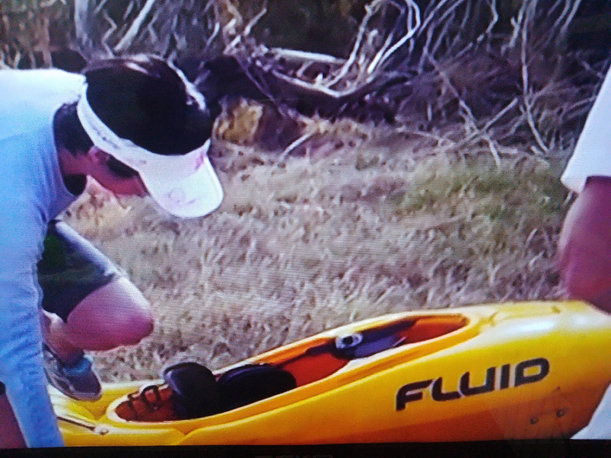 The image shows a person in a blue shirt and a white visor, wearing sunglasses, bending over a yellow kayak. The kayak has the word "FLUID" printed on its side in black letters. The person's legs are visible, and they are wearing dark shorts and sneakers. The kayak appears to be on the ground, which is covered in dry grass and brush. The background is out of focus but suggests an outdoor, possibly natural, setting. There are no recognizable landmarks or elements that would indicate the location as Dushanbe, Tajikistan.