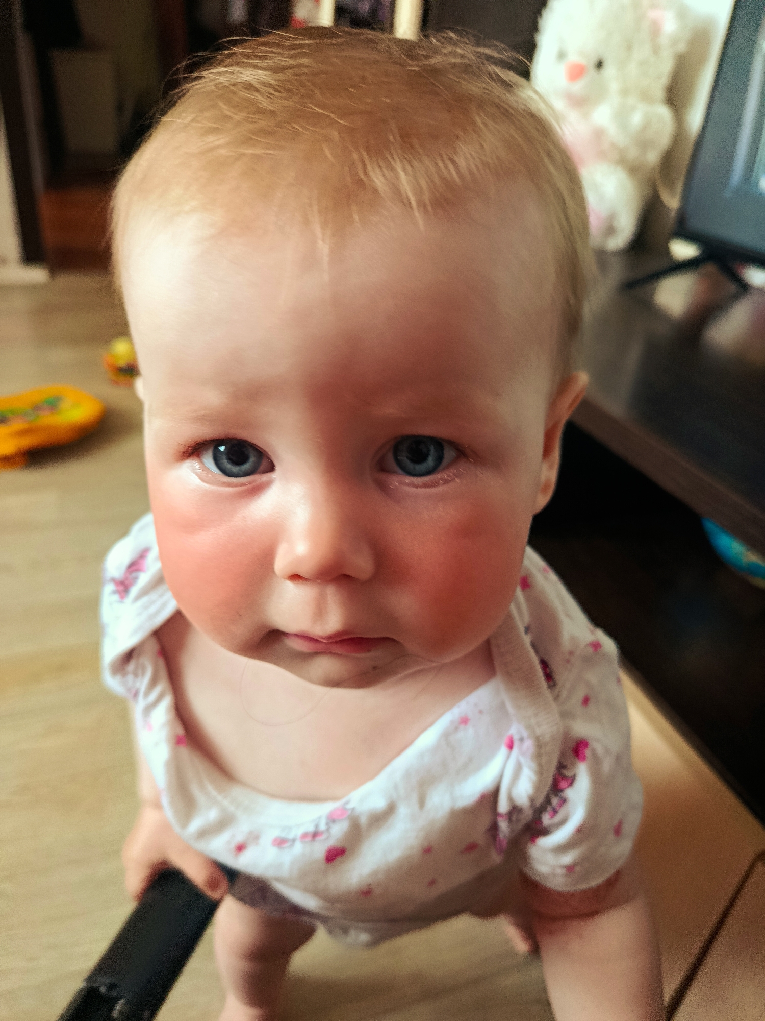 A close-up, high-angle shot depicts a baby with fair skin, light blonde hair, and blue eyes. The baby's cheeks are noticeably red. The child is wearing a white onesie with a pattern of small pink hearts. The baby is holding a black object. In the background, slightly out of focus, are objects including a brown wooden entertainment center, a white stuffed toy, and colorful children's toys on a light-colored floor. The geographical location is not discernible.