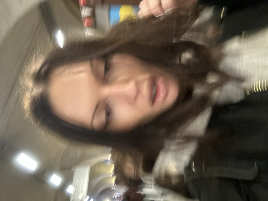 A close-up, motion-blurred image captures a female individual from the chest up, positioned in the foreground of a large indoor public space. The female has long, dark brown hair with a middle part, and her face is angled downwards and slightly to the right, with her mouth partially open. She wears a dark jacket over a light grey hooded garment. The background is significantly blurred but reveals a high, arched ceiling structure with rectangular overhead lighting fixtures. Distant, indistinct figures suggest a crowded environment. Visible on the right side of the background are blurred red and white banners or decorations, and a blue and yellow object, possibly a flag or sign. The architectural style, particularly the vaulted ceiling and lighting, indicates the Western Concourse of King's Cross Station in London, United Kingdom.