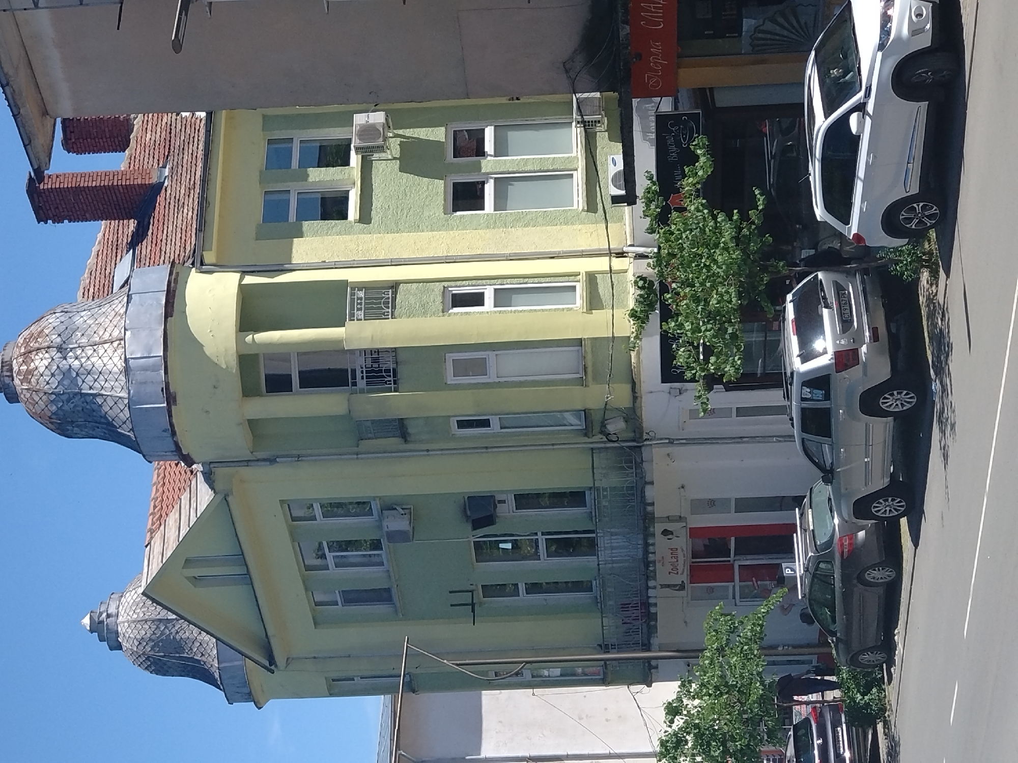 The image shows a street scene in Stara Zagora, Bulgaria, featuring a multi-story building with distinctive architectural elements, including two domed towers. The building is painted in varying shades of green and yellow. It also has a red tiled roof. The building has multiple windows, and air conditioning units are visible on the façade. The ground floor of the building appears to house commercial establishments, including a store with the name "Zooland" visible on the storefront. Cars are parked along the street in front of the building. There are trees planted along the sidewalk. One person is visible walking along the street.
