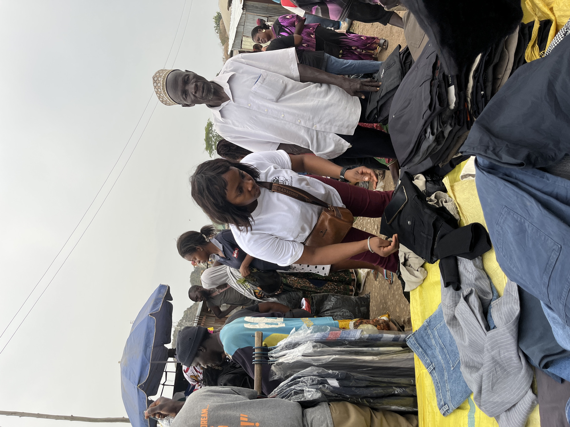 People are browsing clothing on display at an outdoor market in Gura Top, Nigeria. An older man in a white shirt and beaded cap stands near a woman in a white t-shirt and burgundy pants, who is looking at the clothes on a table. Other people are also browsing the goods. Blue umbrellas are visible in the background. The clothing appears to be used.
